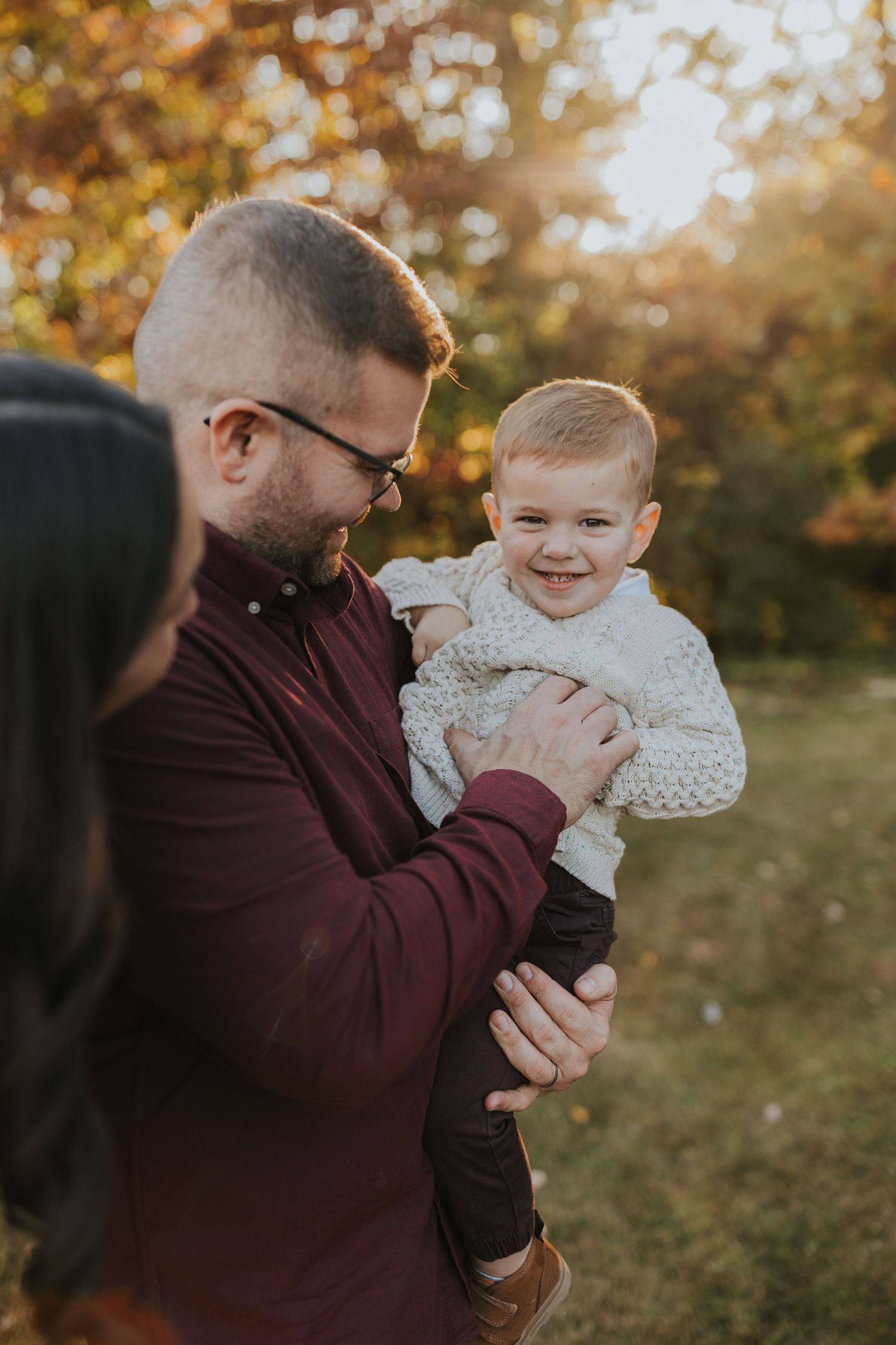 Laughing toddler in cream knit sweater held by father in burgundy shirt during golden hour fall photos.