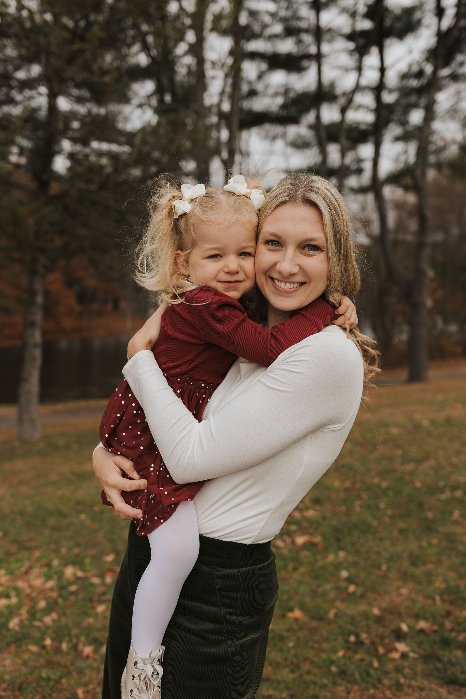Smiling blonde mother holding her young daughter in a red dress during autumn park photos.