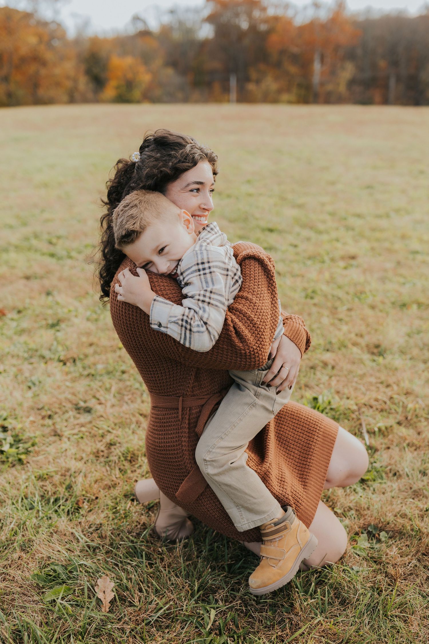 A mother kneels in a grassy field hugging her young son during an autumn family photo session.