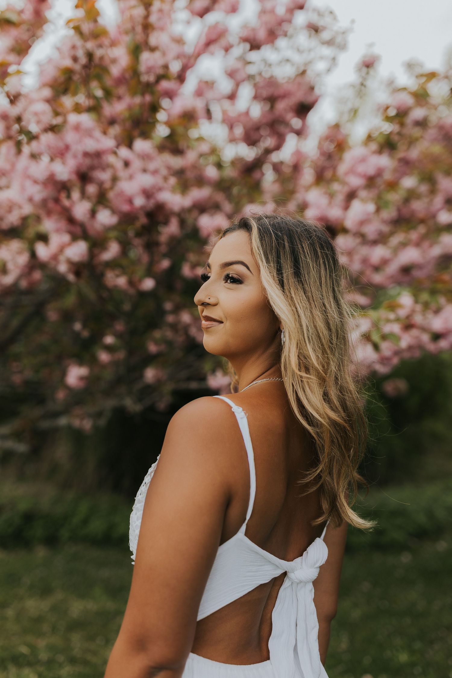 Woman in white backless dress posing before pink cherry blossom trees.