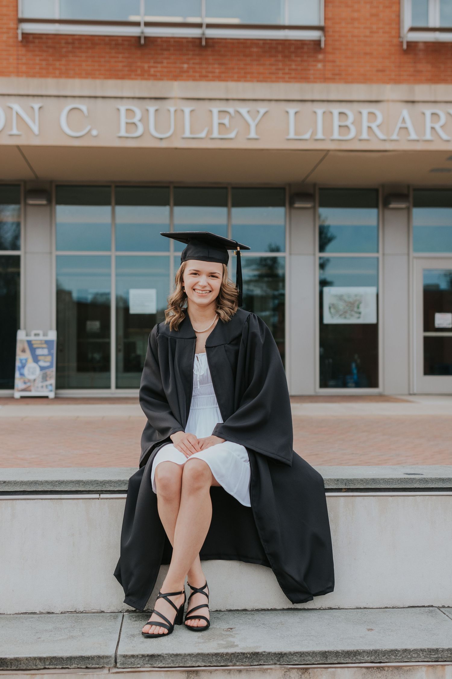 Female graduate in cap and gown sits smiling outside Hilton C. Buley Library.