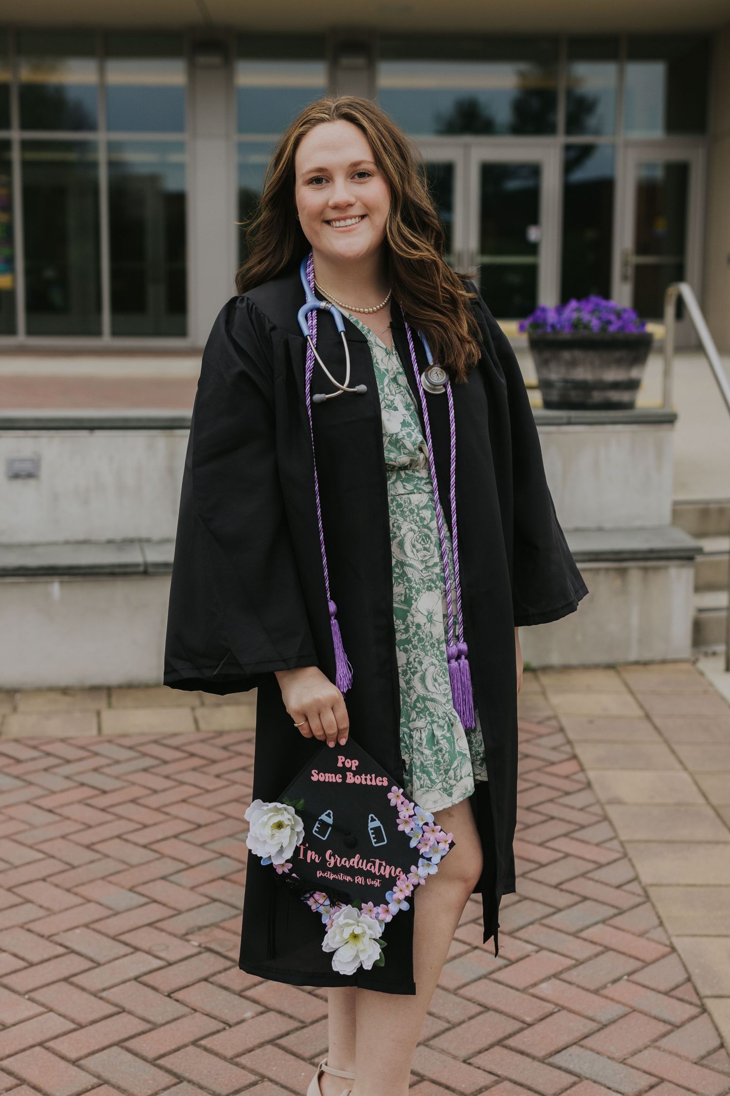 Nursing graduate in black gown holding decorated cap with stethoscope outside library.