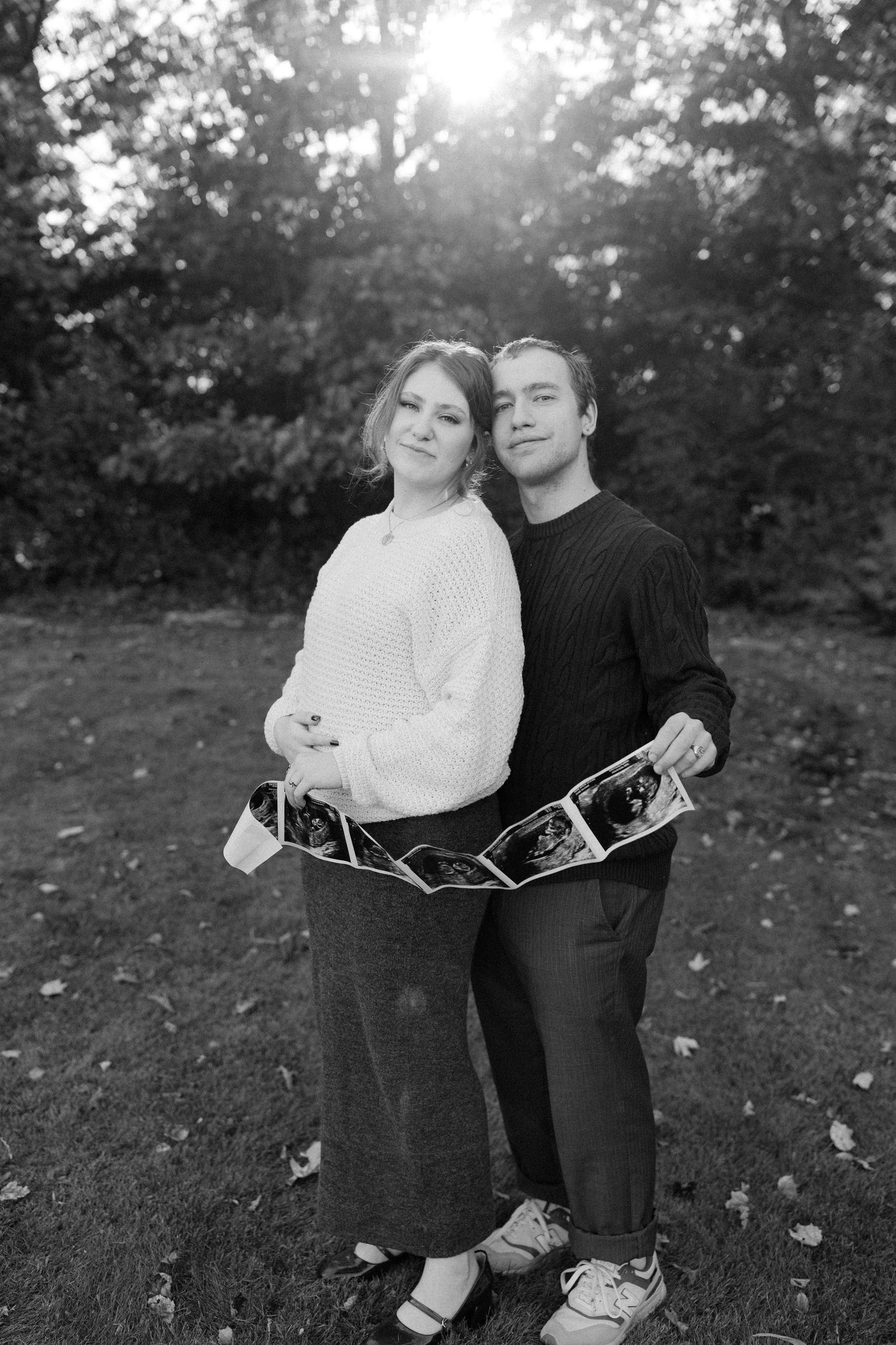 Black and white photo of couple embracing while holding ultrasound strip during outdoor maternity photo session.