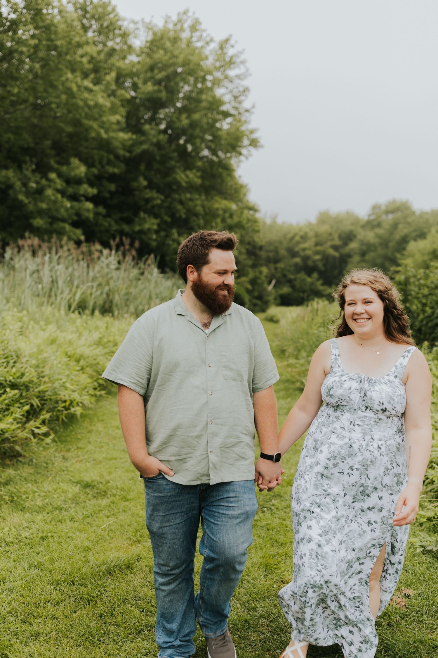 Smiling couple walking hand in hand through a green meadow, man in sage shirt and jeans, woman in floral dress.