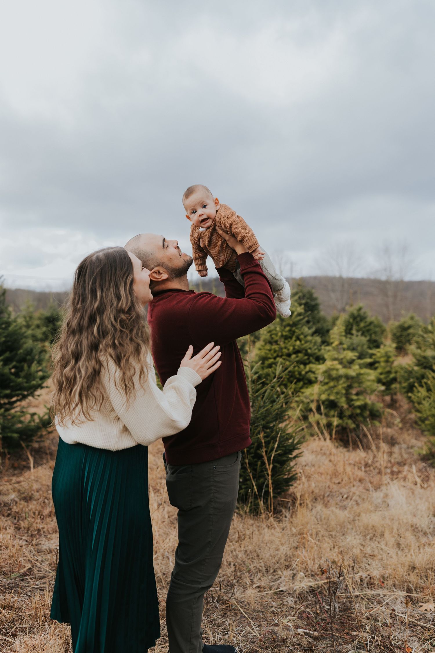 A couple holds their baby up at a Christmas tree farm, wearing festive green and burgundy outfits on a cloudy day.