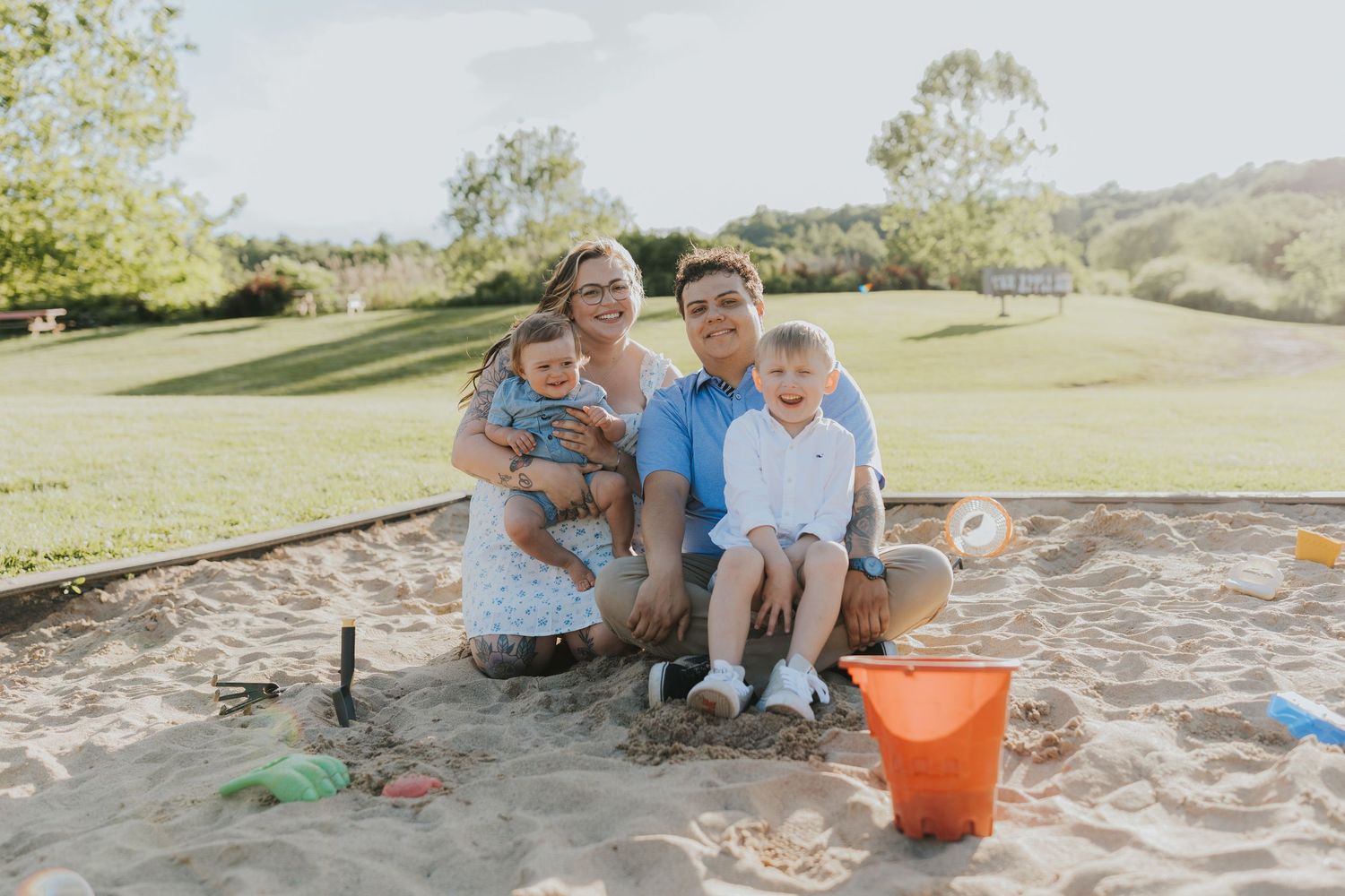 Family of four smiling together in a sandbox outdoors, with green hills and playground toys visible in the background.