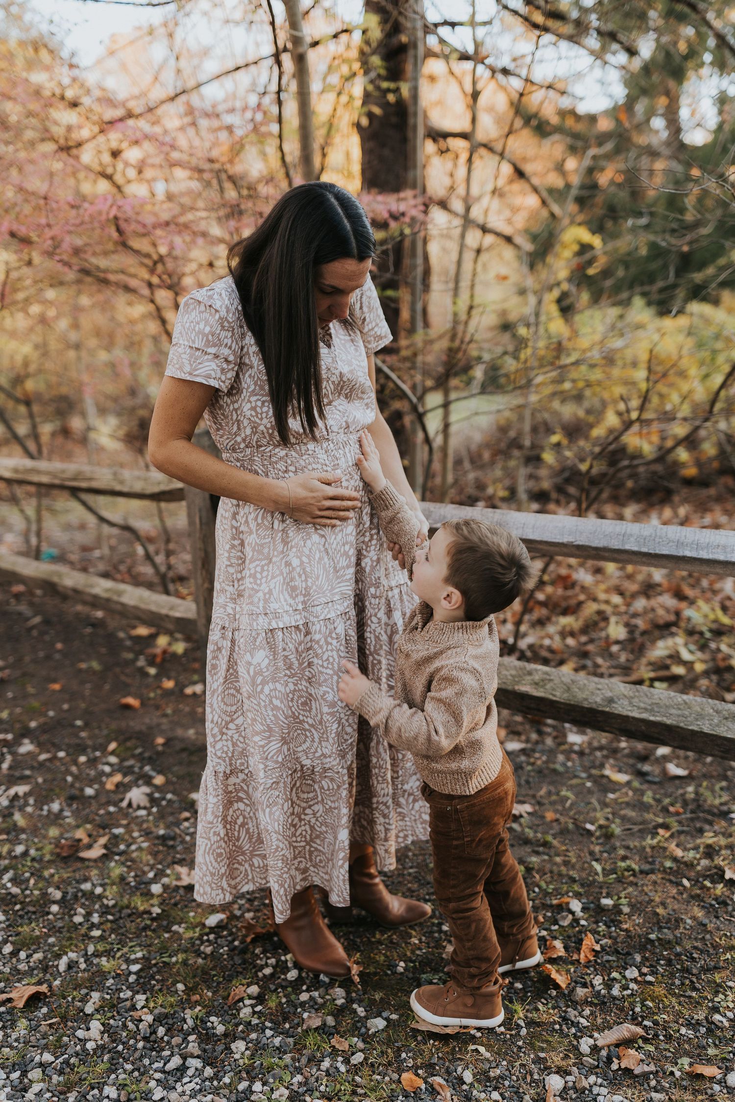 Pregnant woman in floral dress with toddler touching her baby bump outdoors in autumn park.