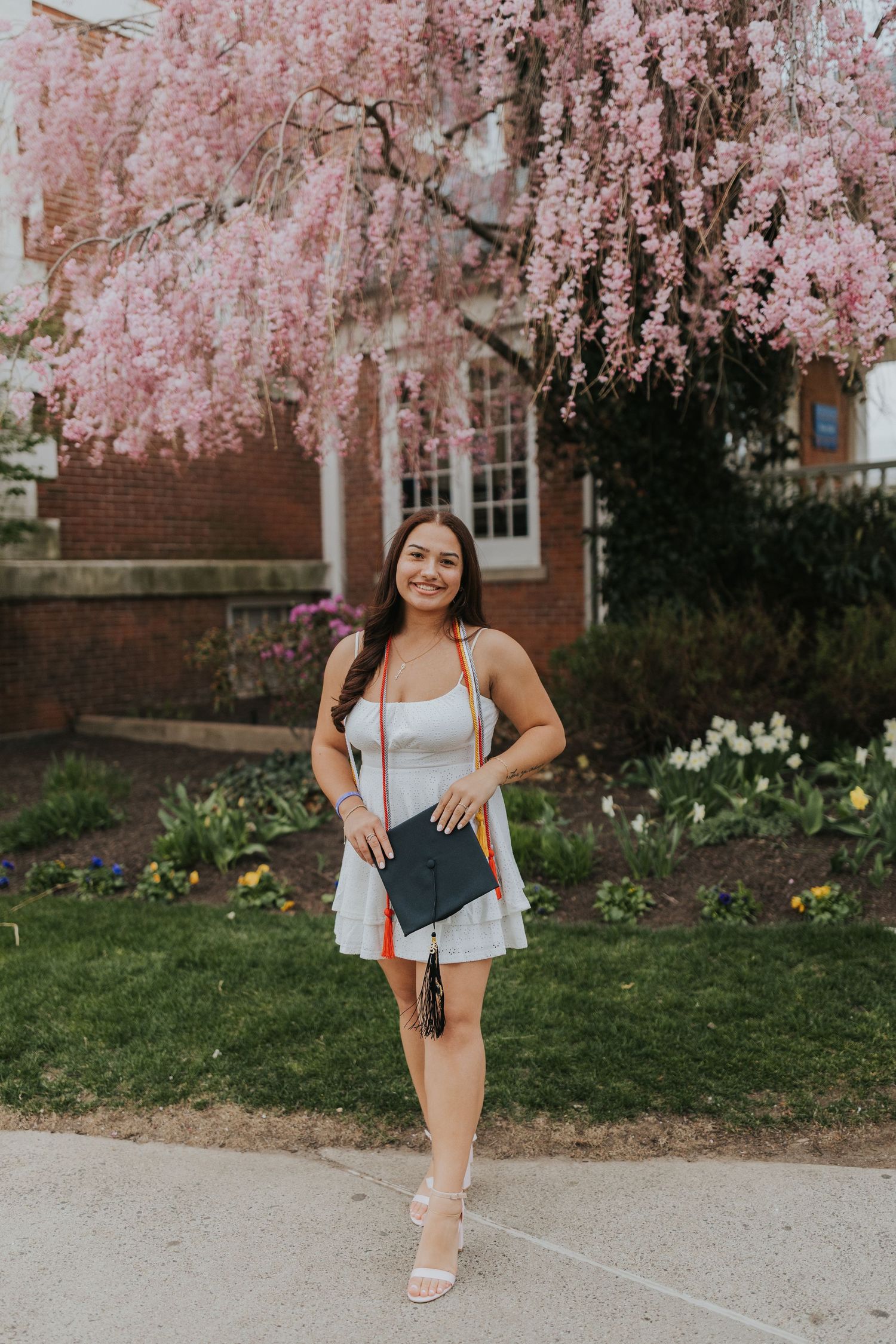 Smiling graduate in white dress holds cap near pink cherry blossom tree on college campus.