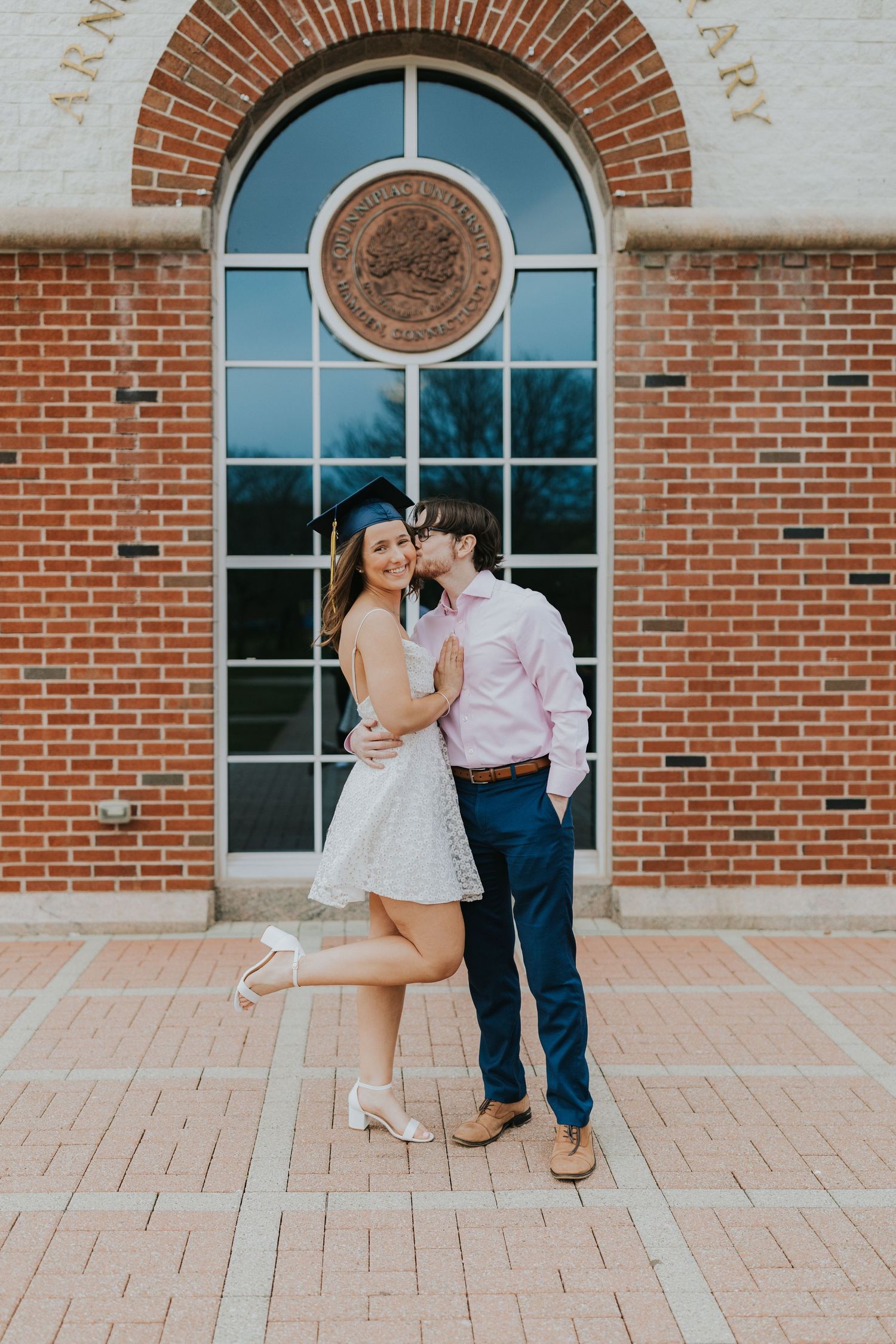 Graduate in white dress and cap receiving a kiss on the cheek outside a brick university building.