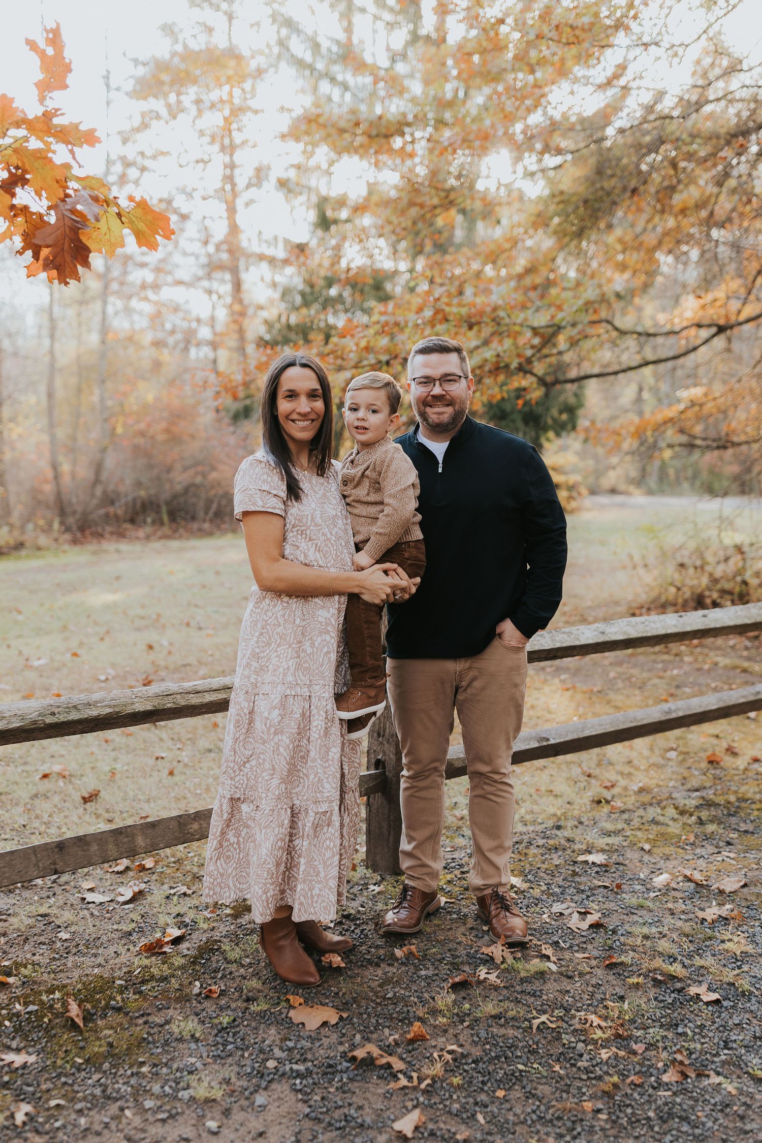 Family of three smiling together outdoors surrounded by vibrant fall foliage and autumn leaves.