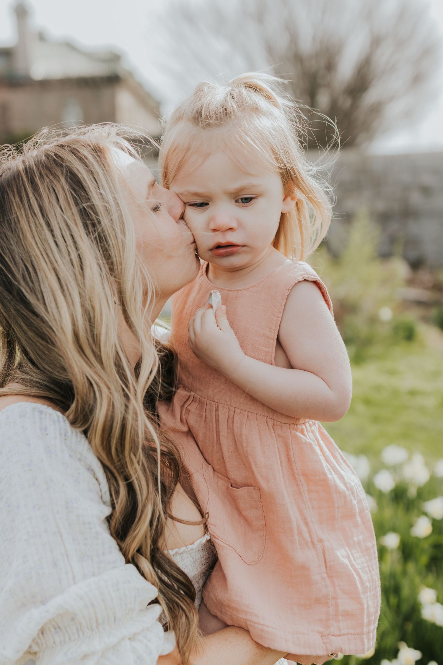 Mother tenderly kisses cheek of blonde toddler girl in pink dress outdoors in spring garden.