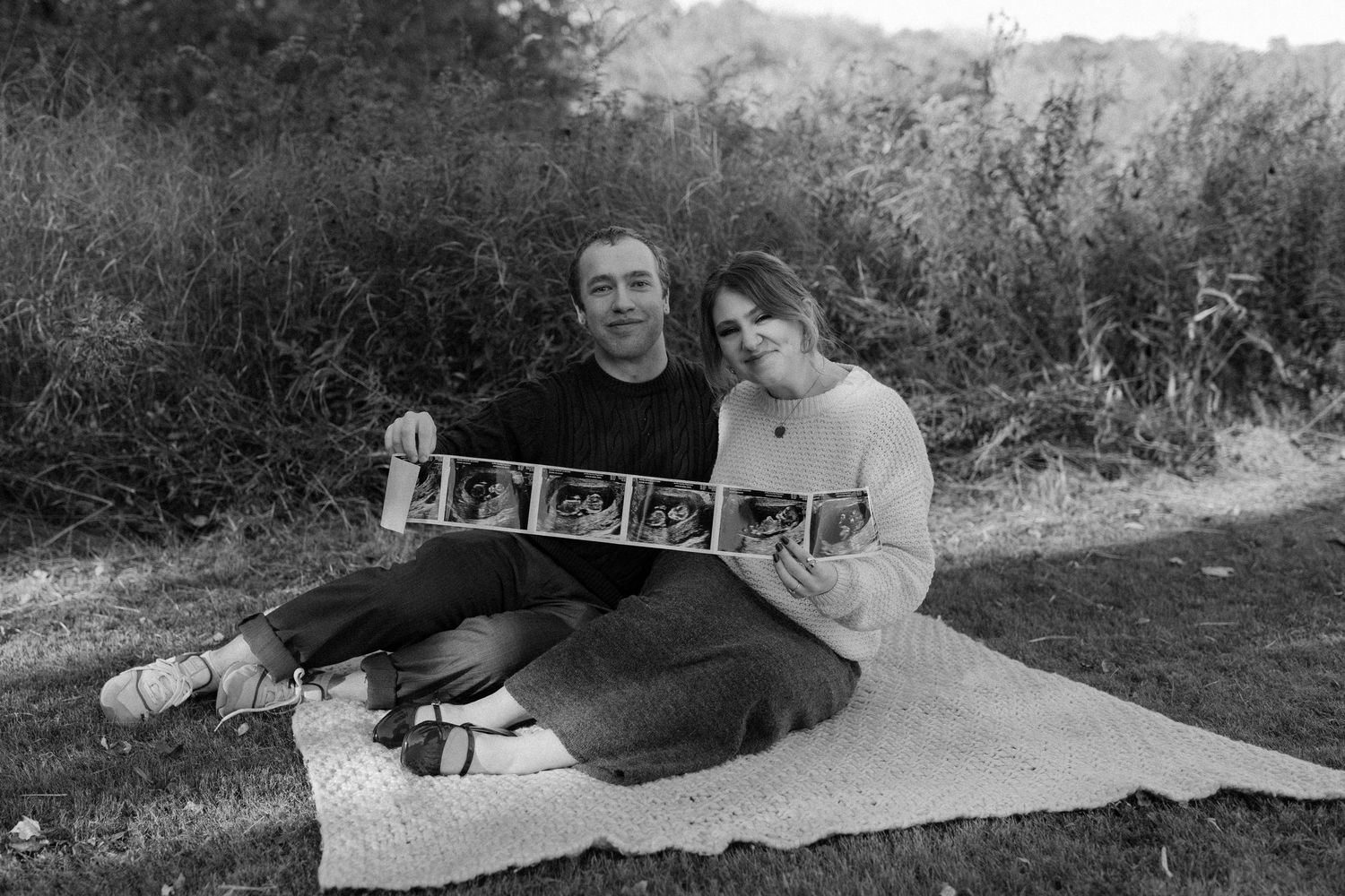 Black and white photo of couple sitting on blanket holding ultrasound strip in natural outdoor setting.