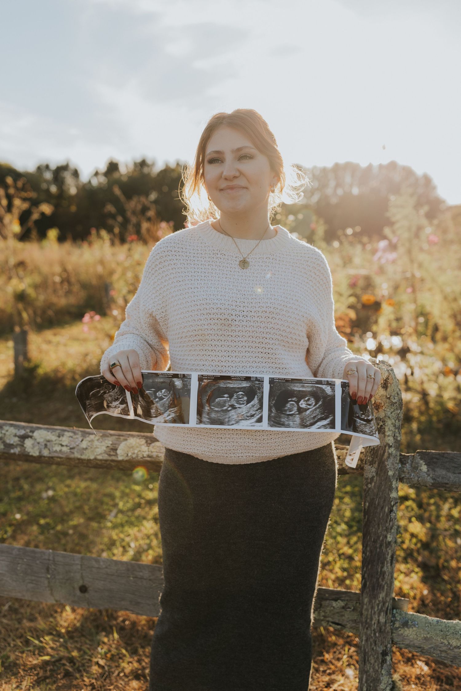 Pregnant woman standing by wooden fence holding ultrasound photos in sunny field, pregnancy announcement.