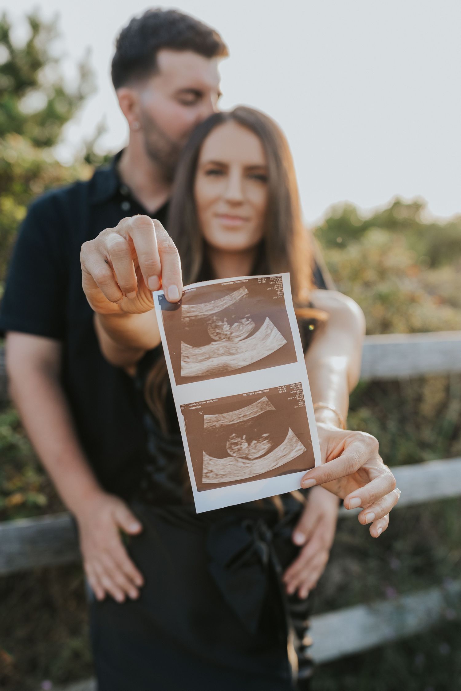 Couple holding ultrasound photos toward camera during golden hour pregnancy announcement photoshoot outdoors.