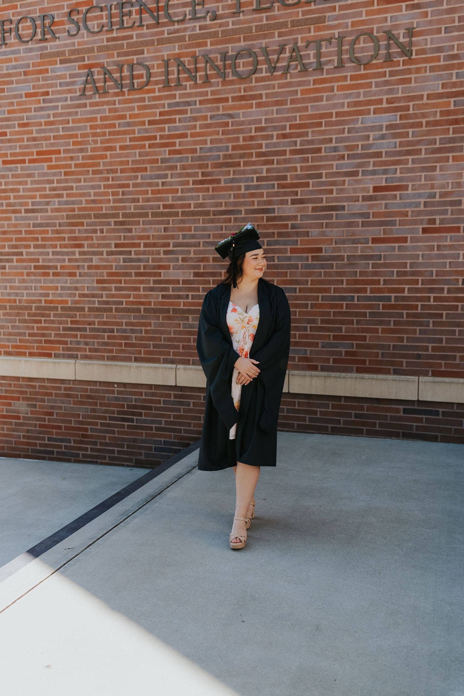 Graduate in black cap and gown walking along brick wall inscribed with Science, Tech and Innovation.