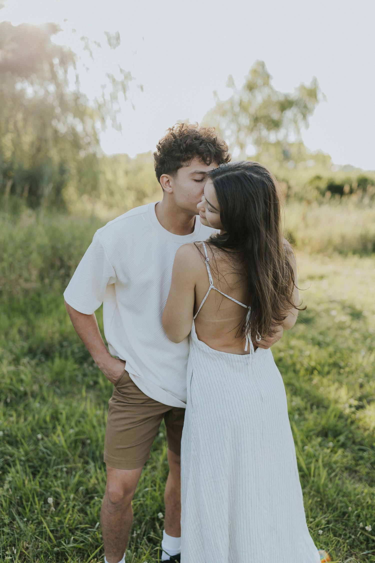 Young couple sharing a tender kiss while standing in a sunlit green meadow outdoors.