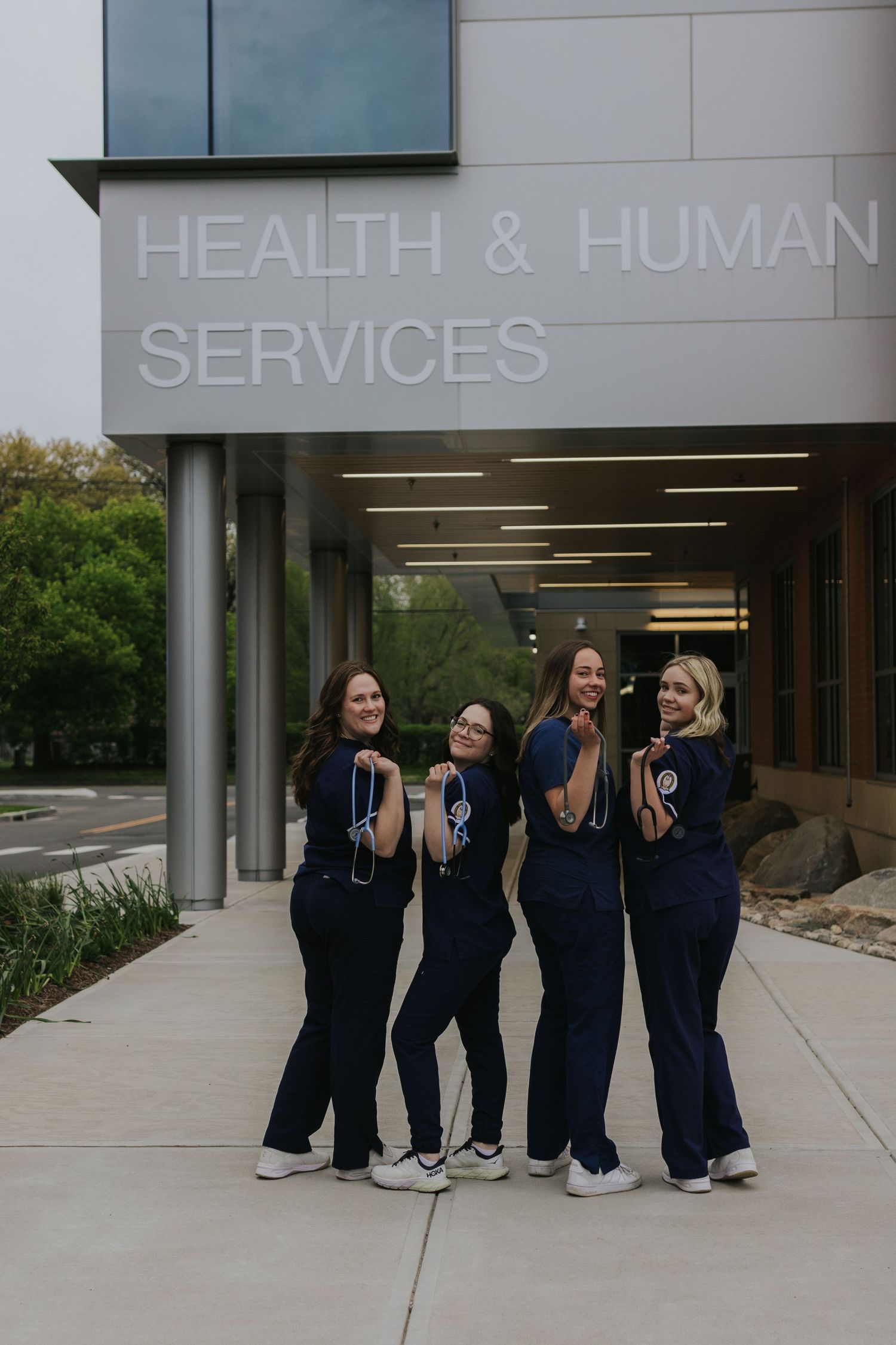 Four nursing students in navy scrubs with stethoscopes outside Health and Human Services building.