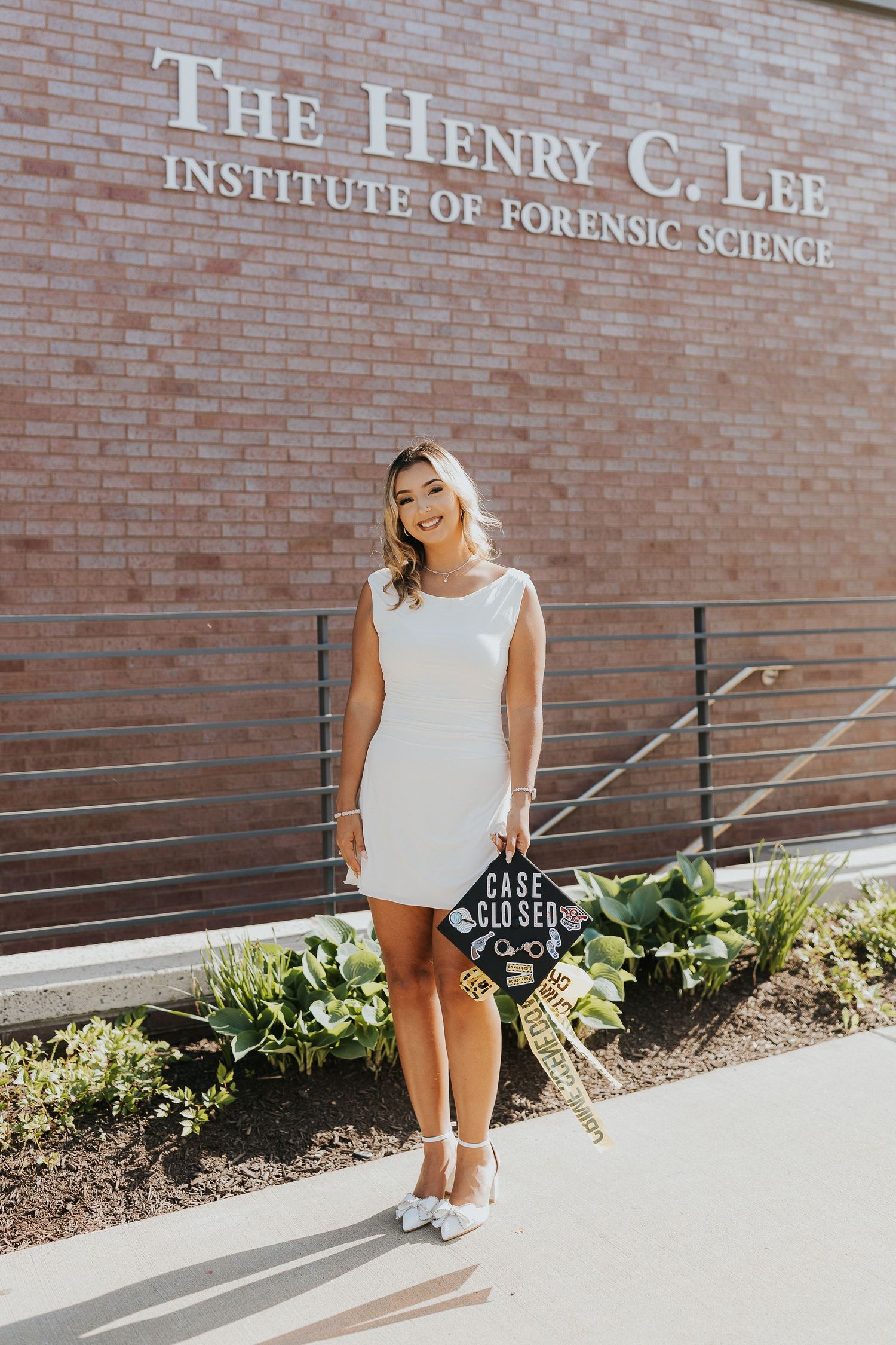 Graduate in white dress stands before The Henry C. Lee Institute of Forensic Science building holding decorated cap.