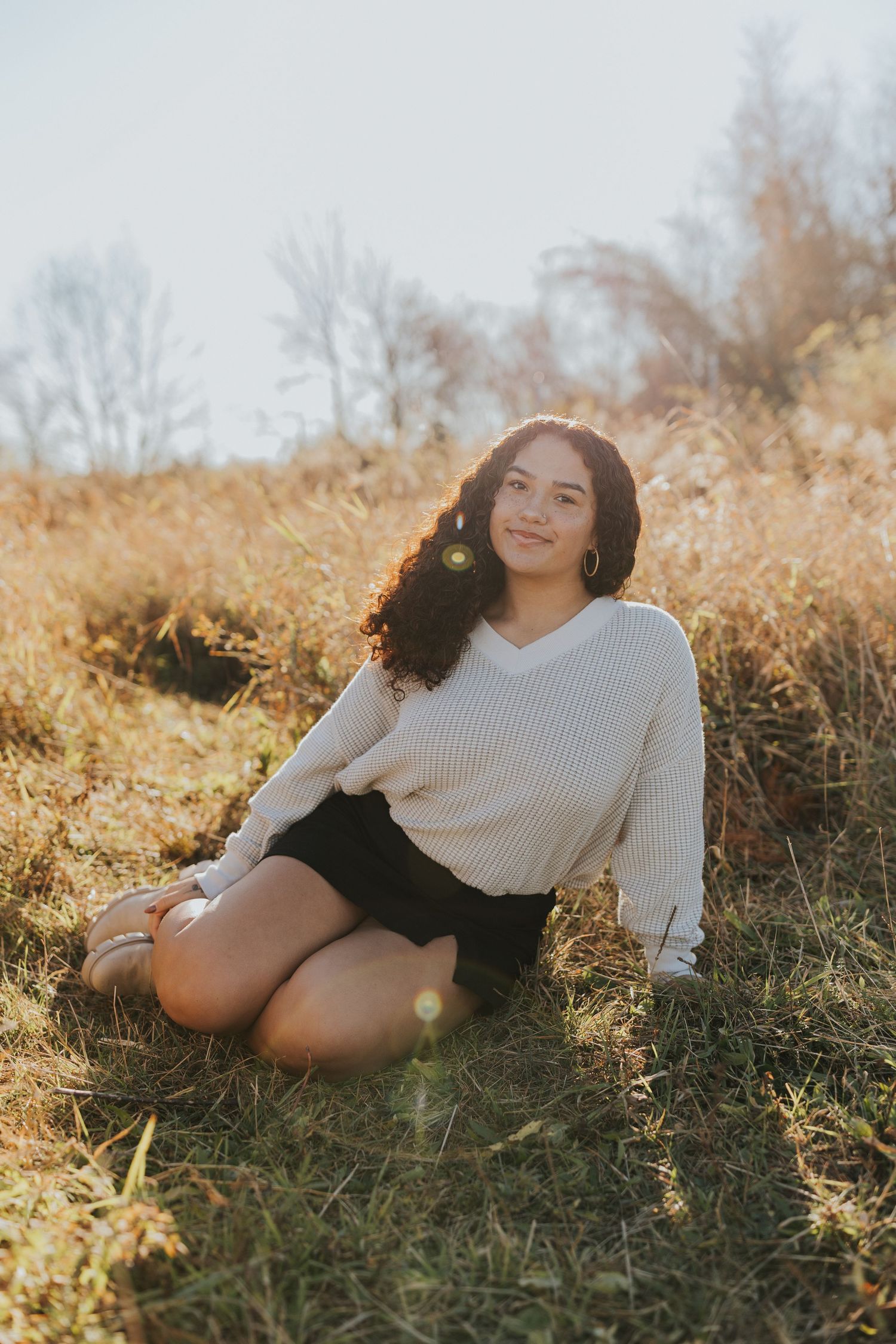 Young woman with curly hair sitting cross-legged in golden autumn field in cream sweater.