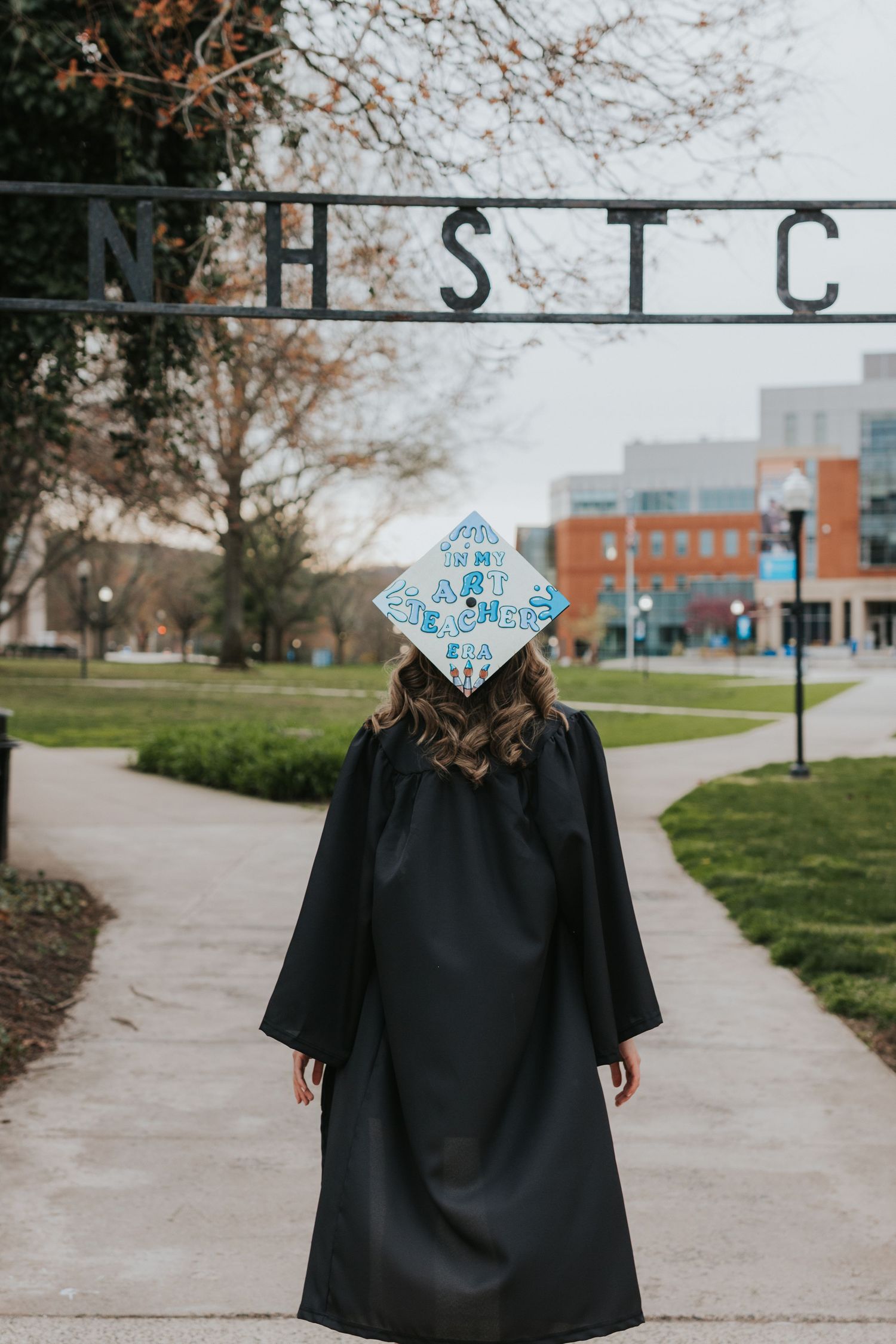 Graduate in cap and gown walks away showing decorated graduation cap under NHSTC gate.