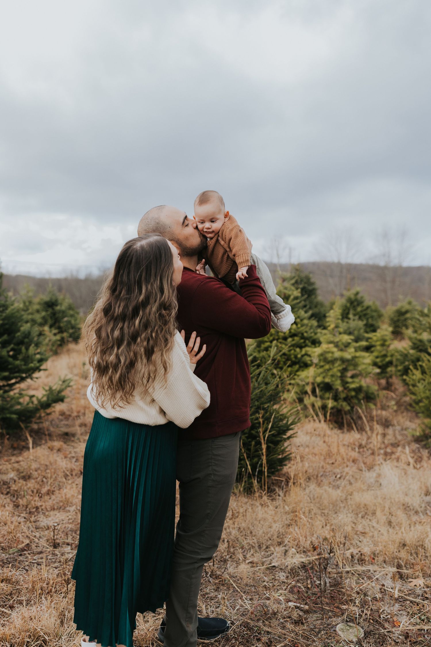Parents at Christmas tree farm, father lifting baby for a kiss while mother stands close.