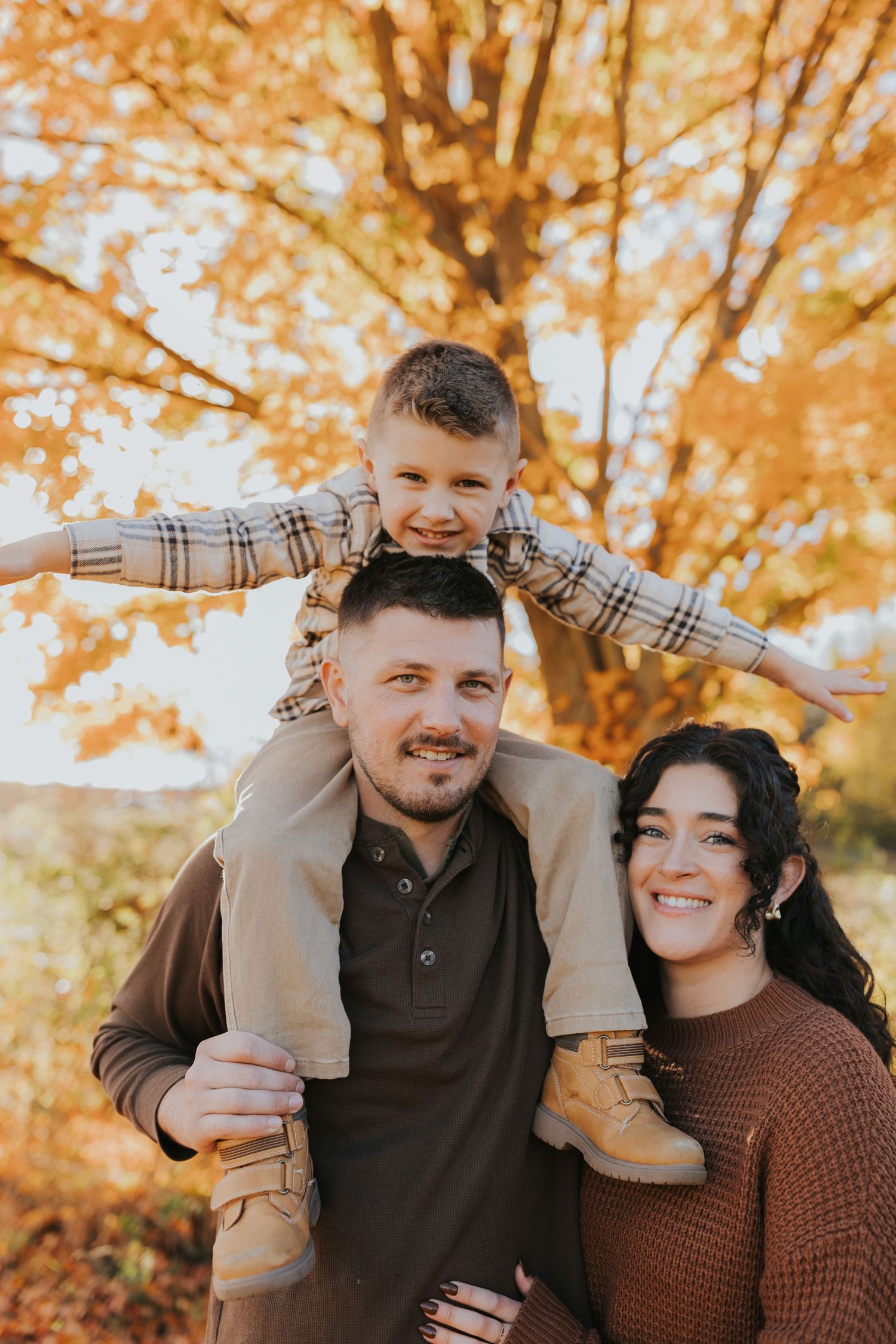 A smiling young boy sits on his father's shoulders while mom stands beside them in fall setting.