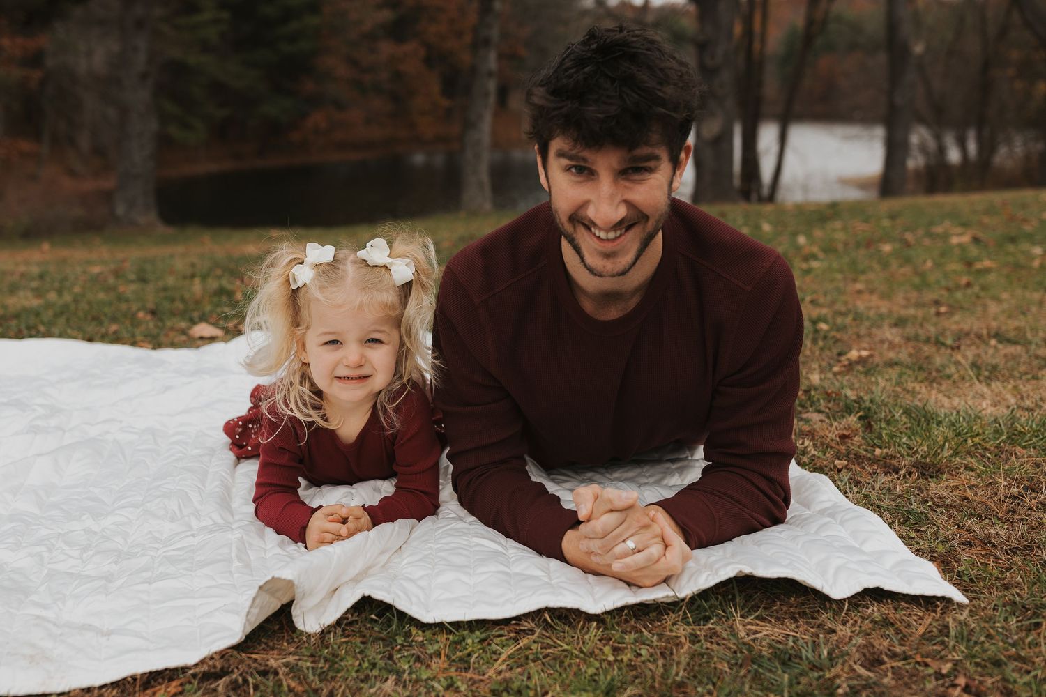 Smiling dark-haired father lying on a white blanket with his young daughter in autumn.