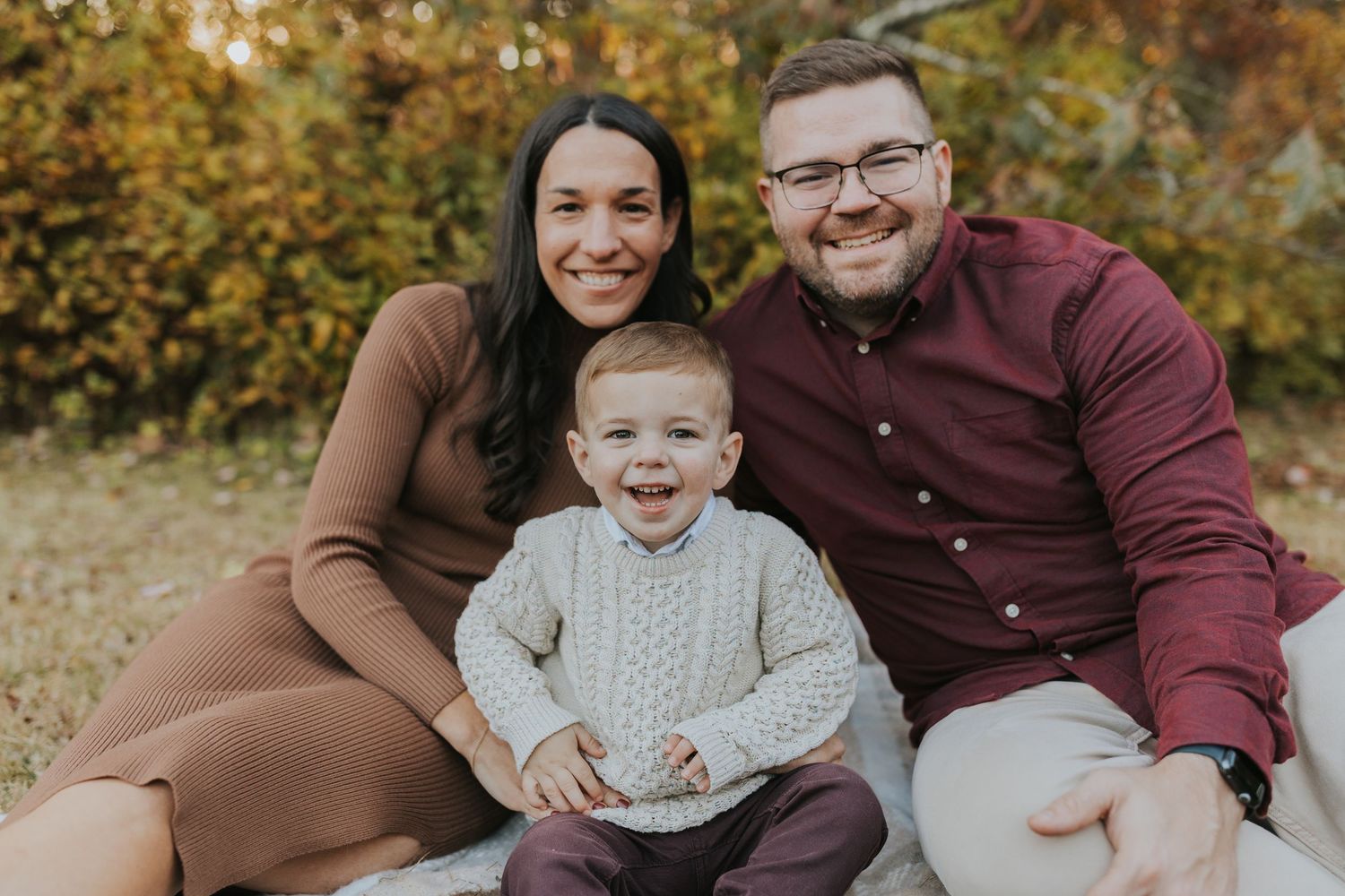 Happy family of three sitting together outdoors smiling during fall mini session in autumn setting.