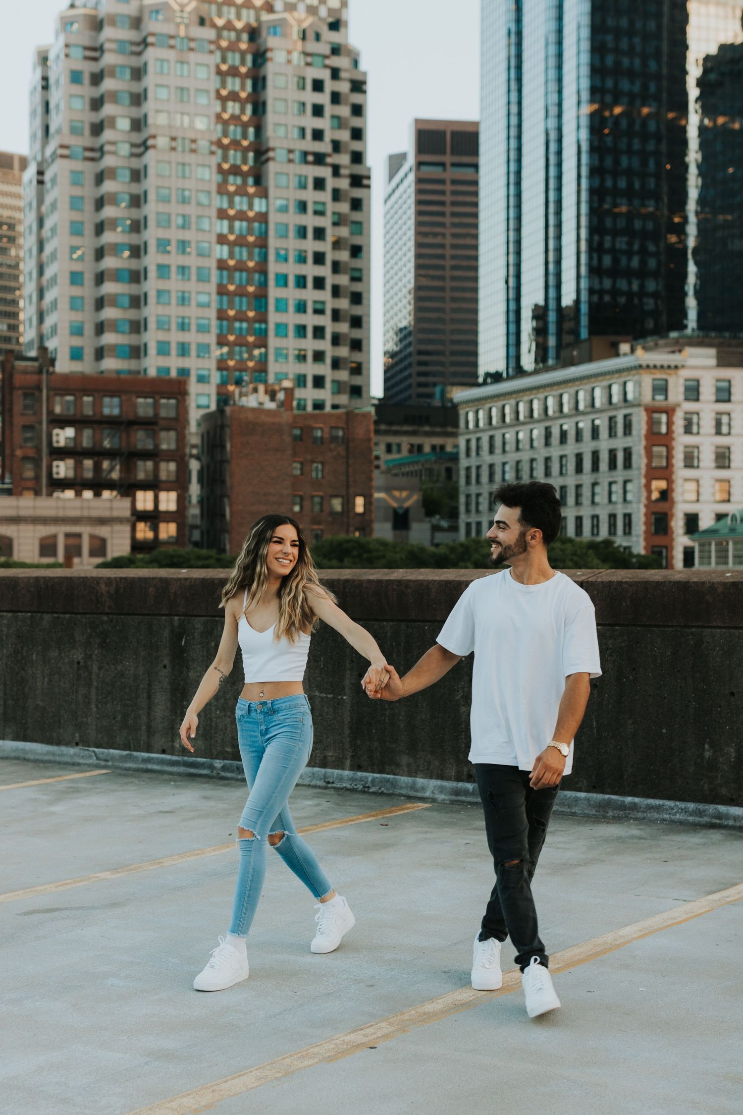 Couple holding hands and walking on a rooftop parking deck with a stunning urban city skyline behind them.