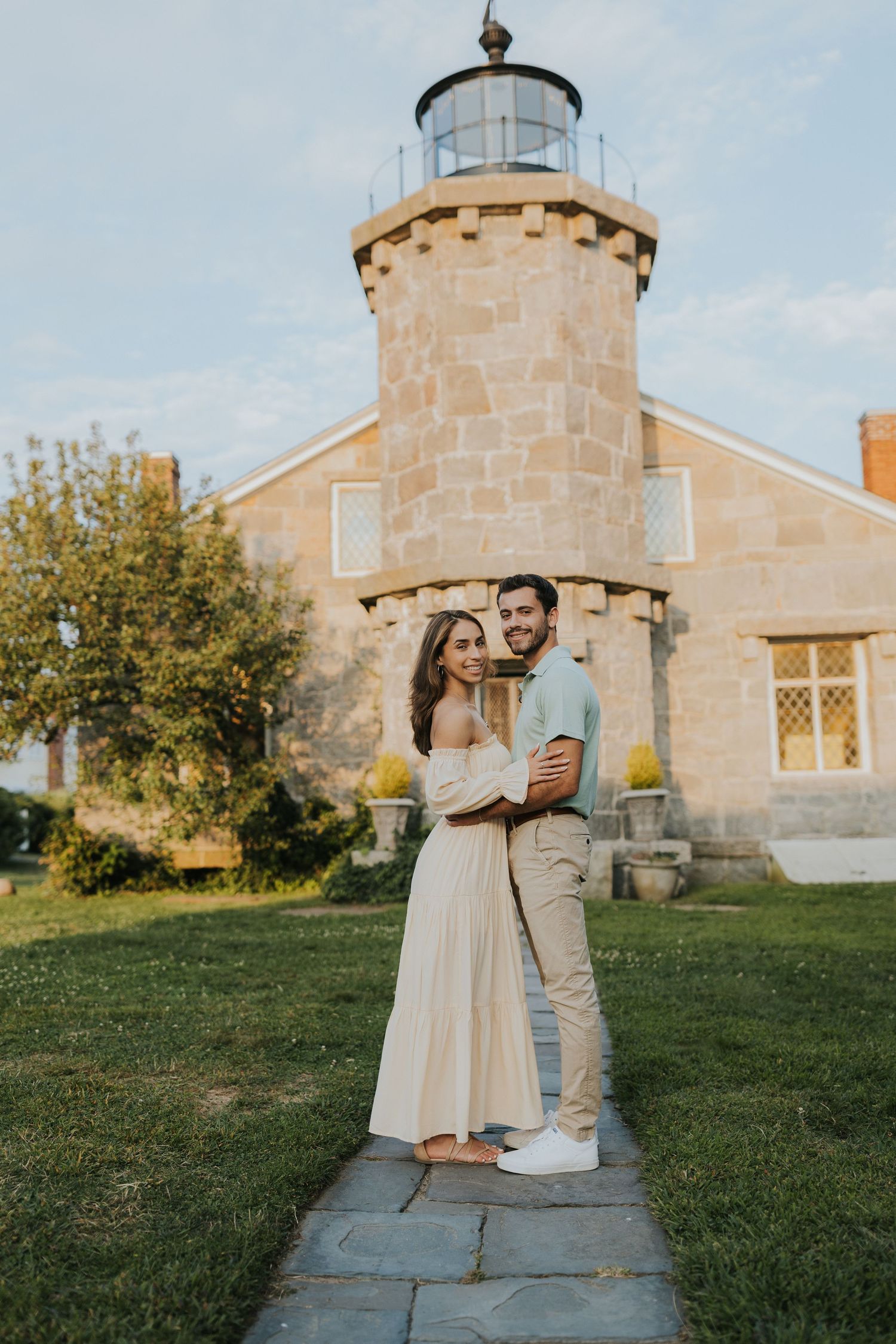 Happy couple embracing in front of historic stone lighthouse on sunny day, woman in cream dress and man in blue polo.