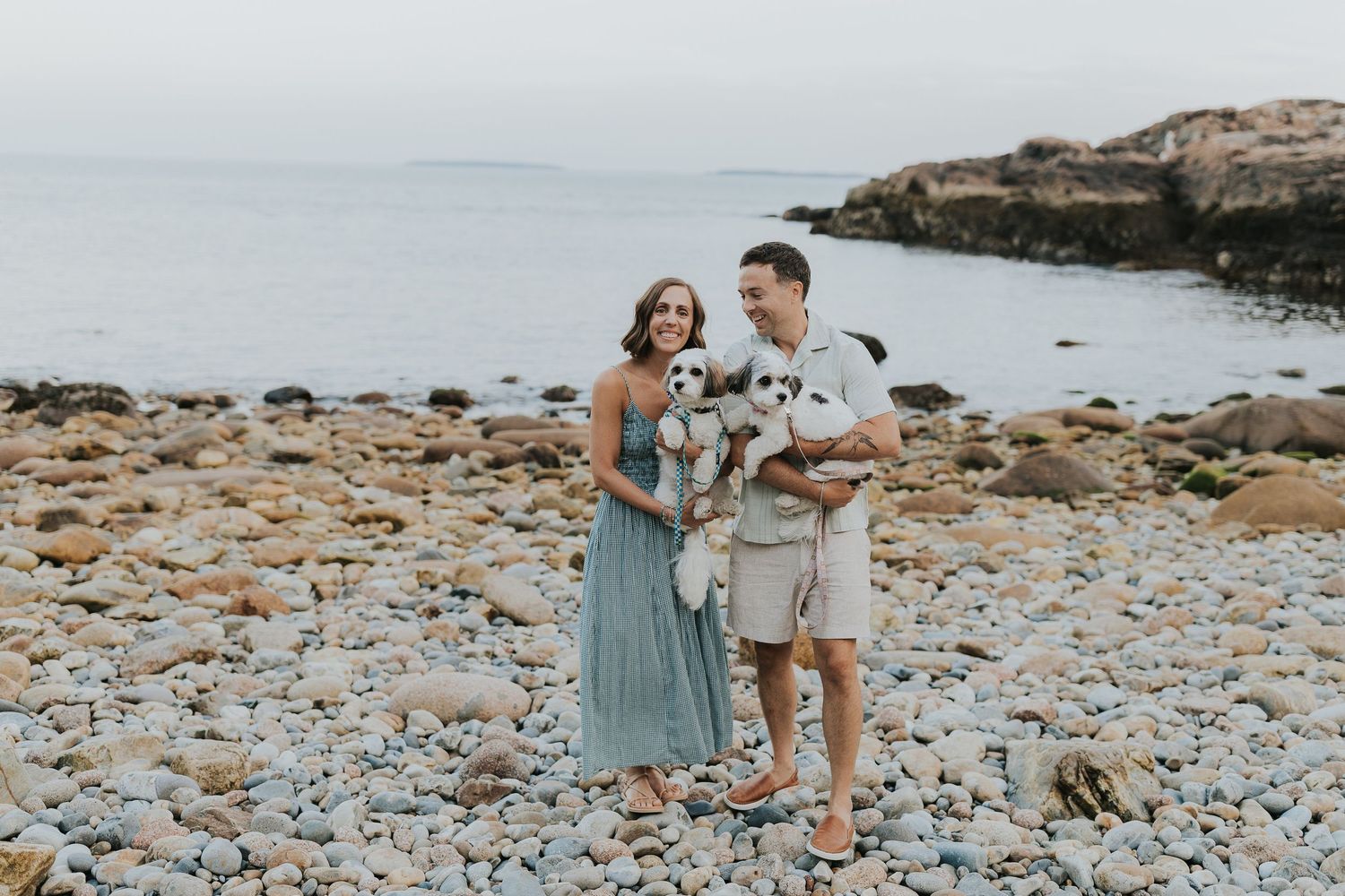 Couple smiling on rocky beach holding two small dogs with ocean in background.