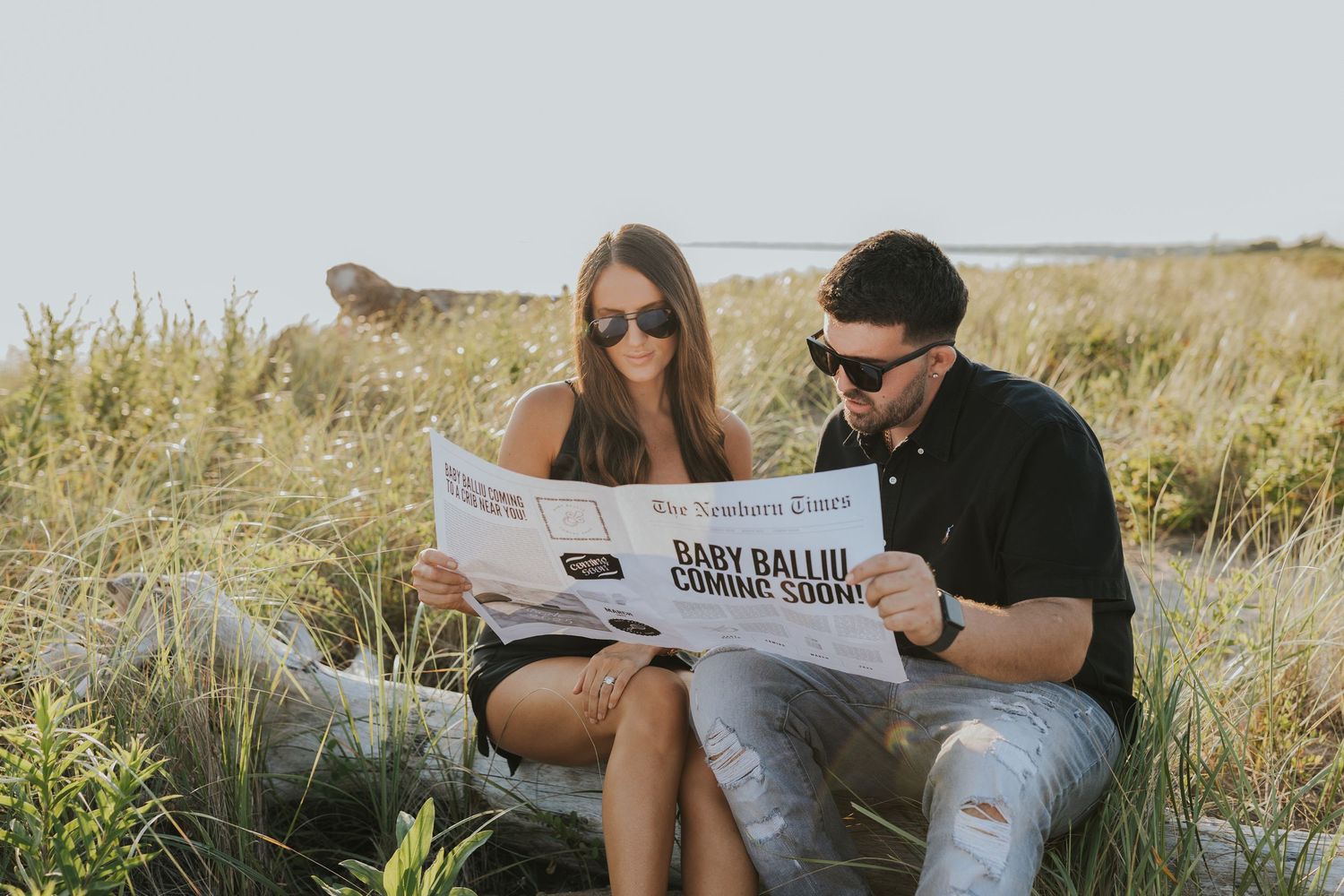 Couple reading a newspaper with Baby Balloon Coming Soon headline while sitting outdoors in a grassy field.