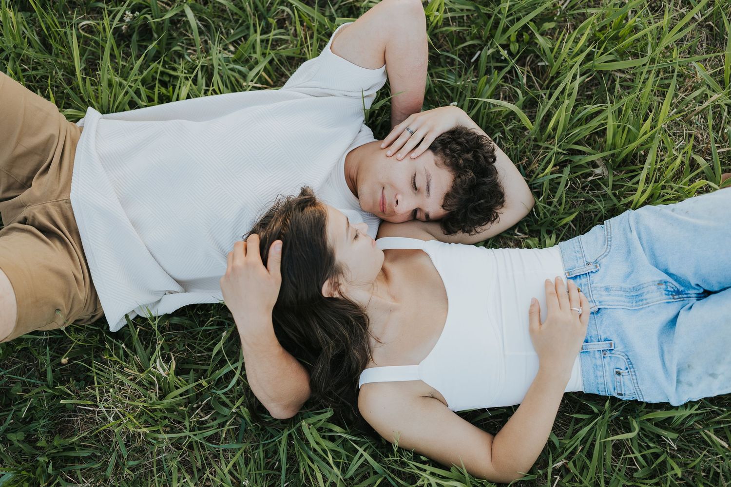 Young couple lies in green grass facing each other in an intimate romantic outdoor portrait.
