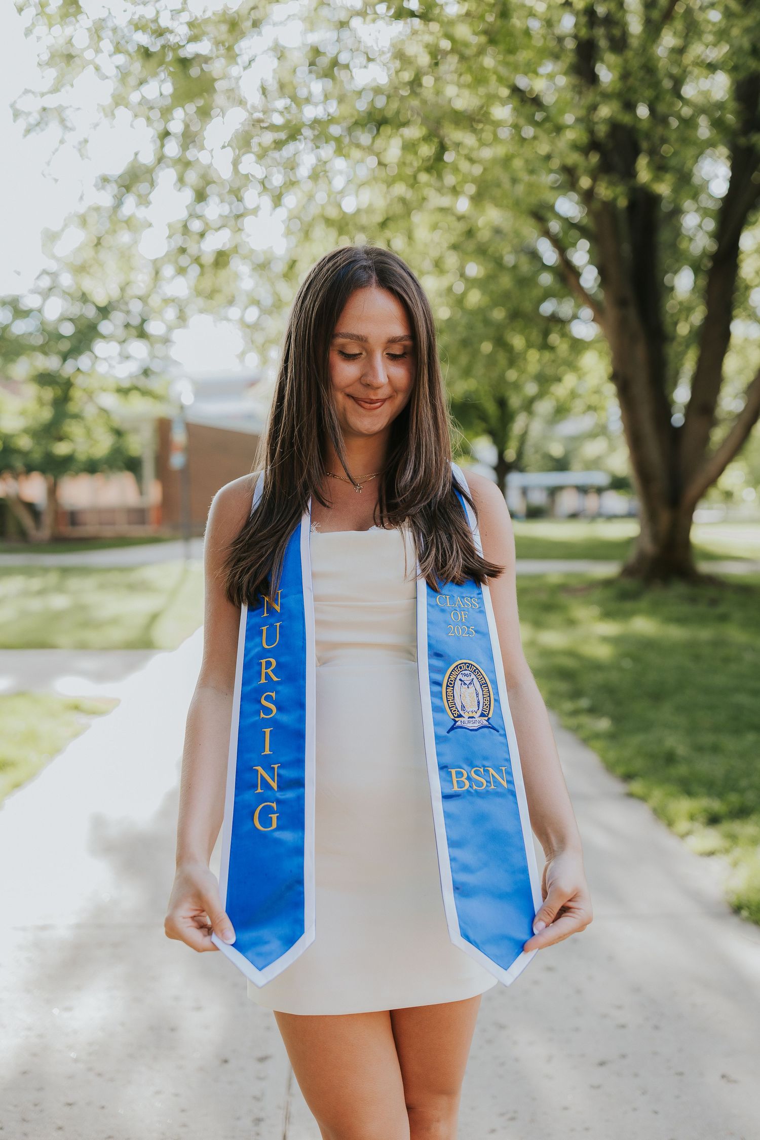 Nursing BSN graduate in white dress displays blue graduation sash outdoors.