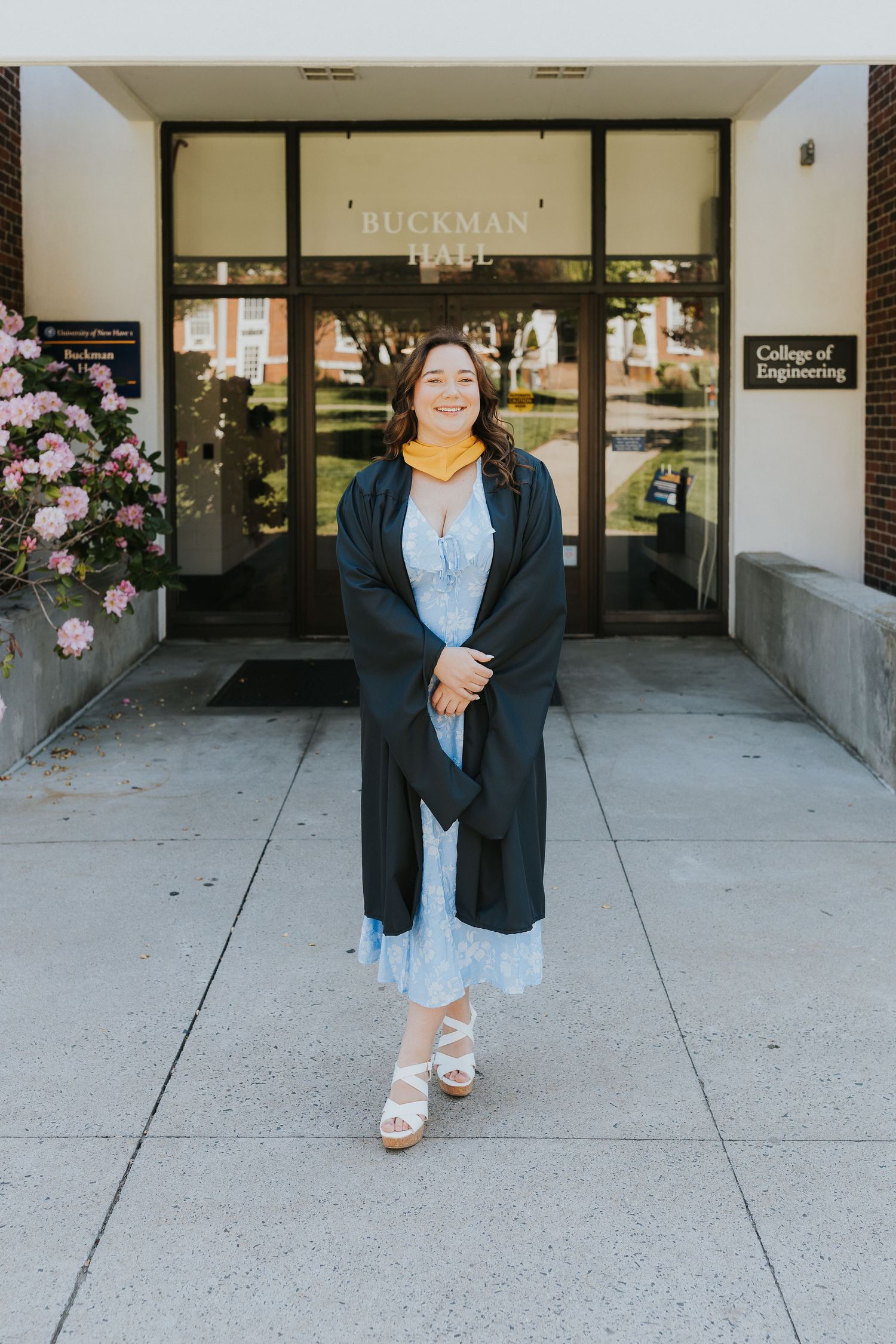 Smiling graduate in black gown and yellow hood standing outside Buckman Hall College of Engineering.