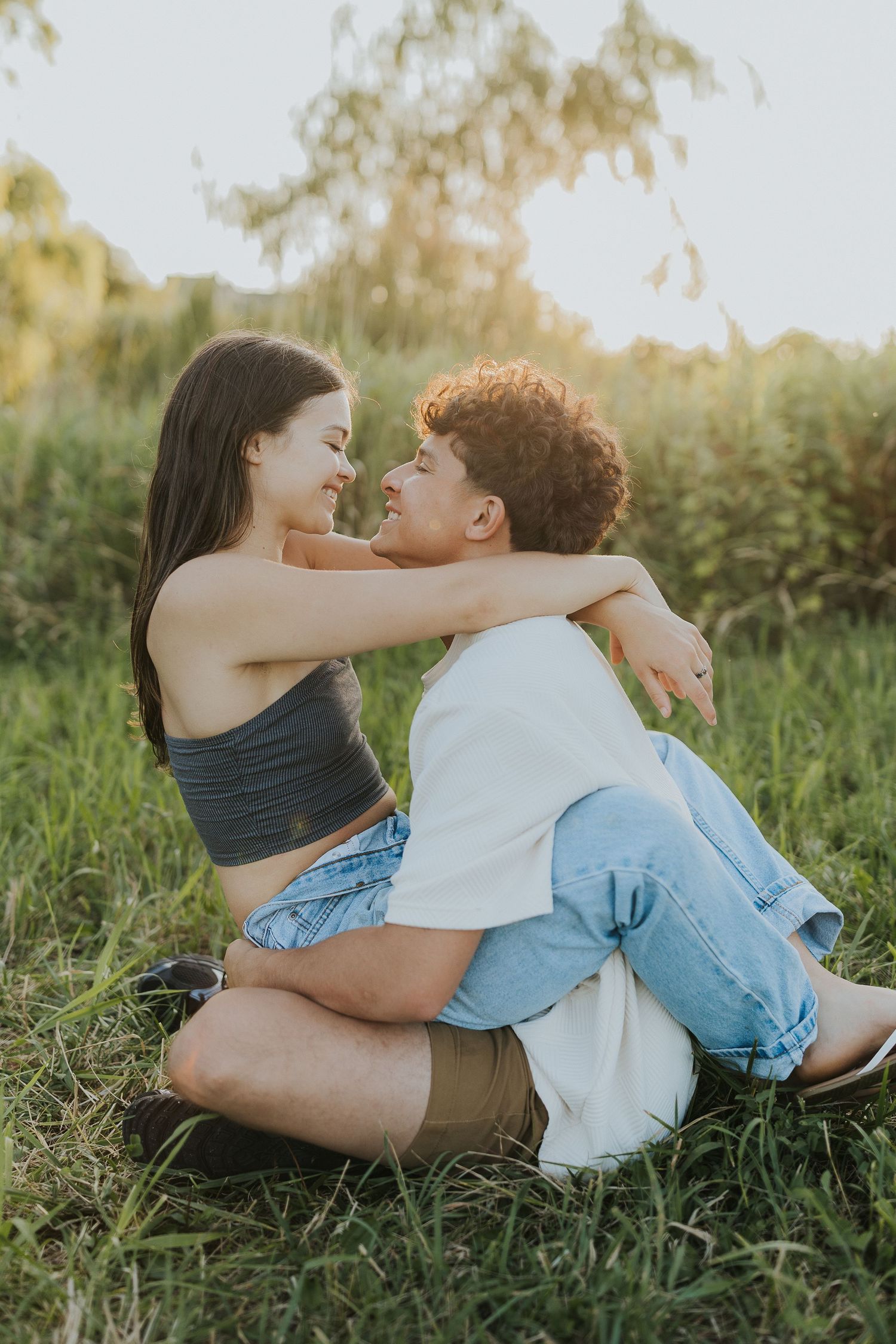 Young couple sitting in grass, embracing and gazing at each other during golden hour outdoors.