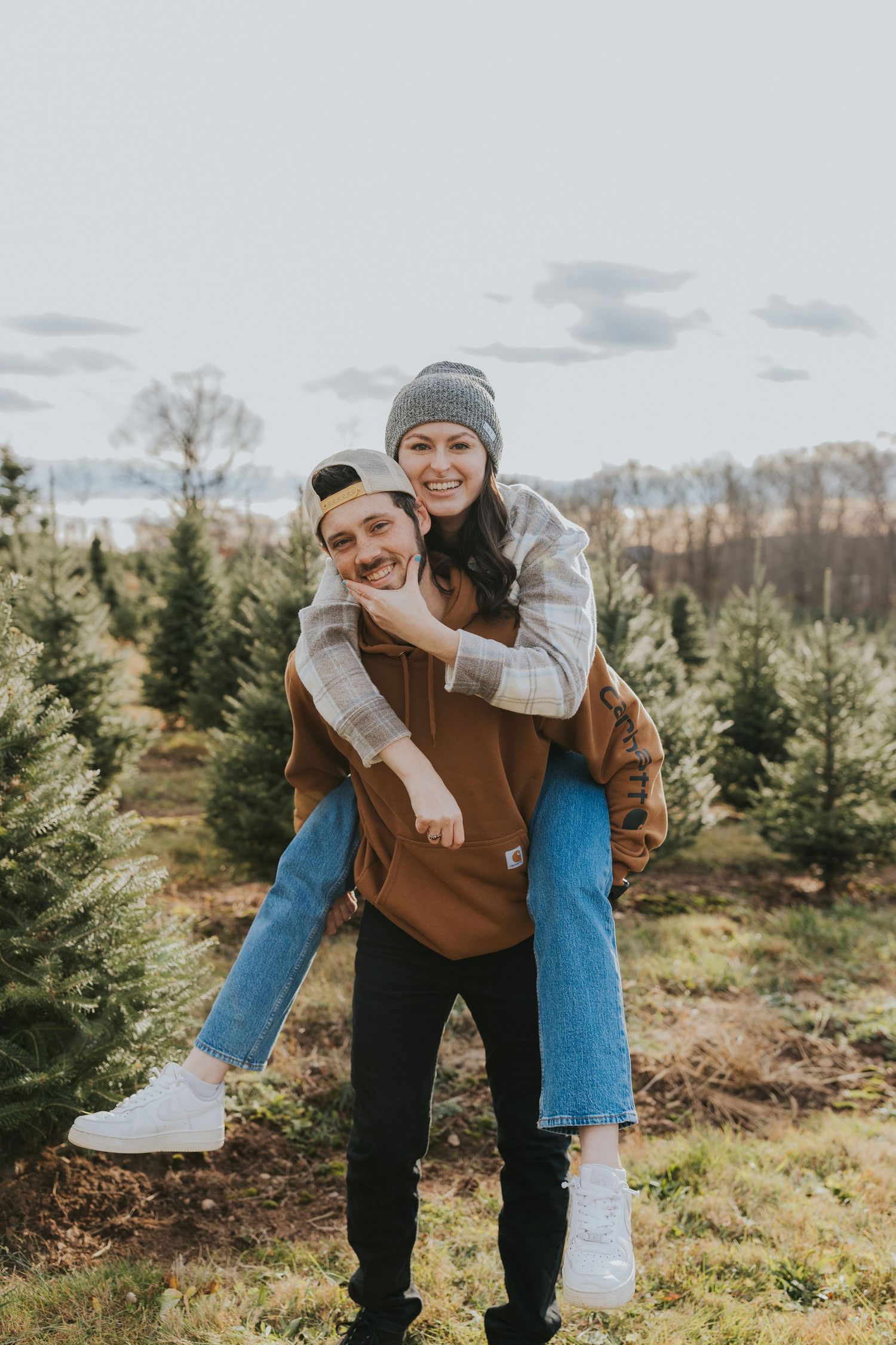 Man giving woman a piggyback ride while smiling at a Christmas tree farm.