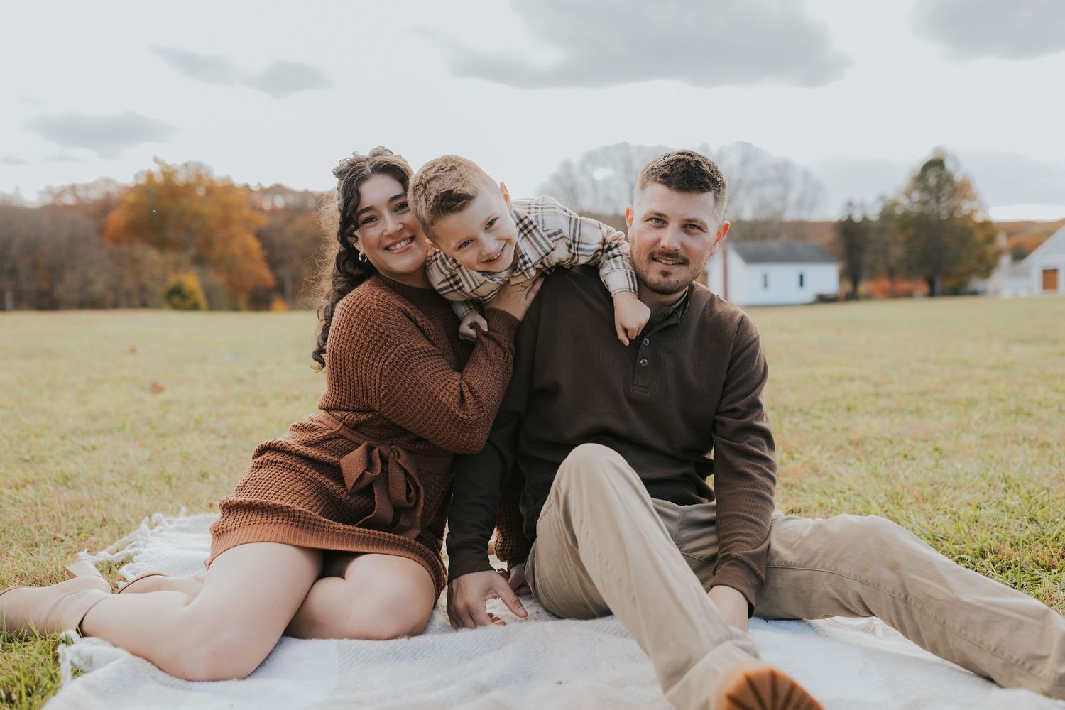 A family of three sits together on a blanket in an open field during a fall photo session.