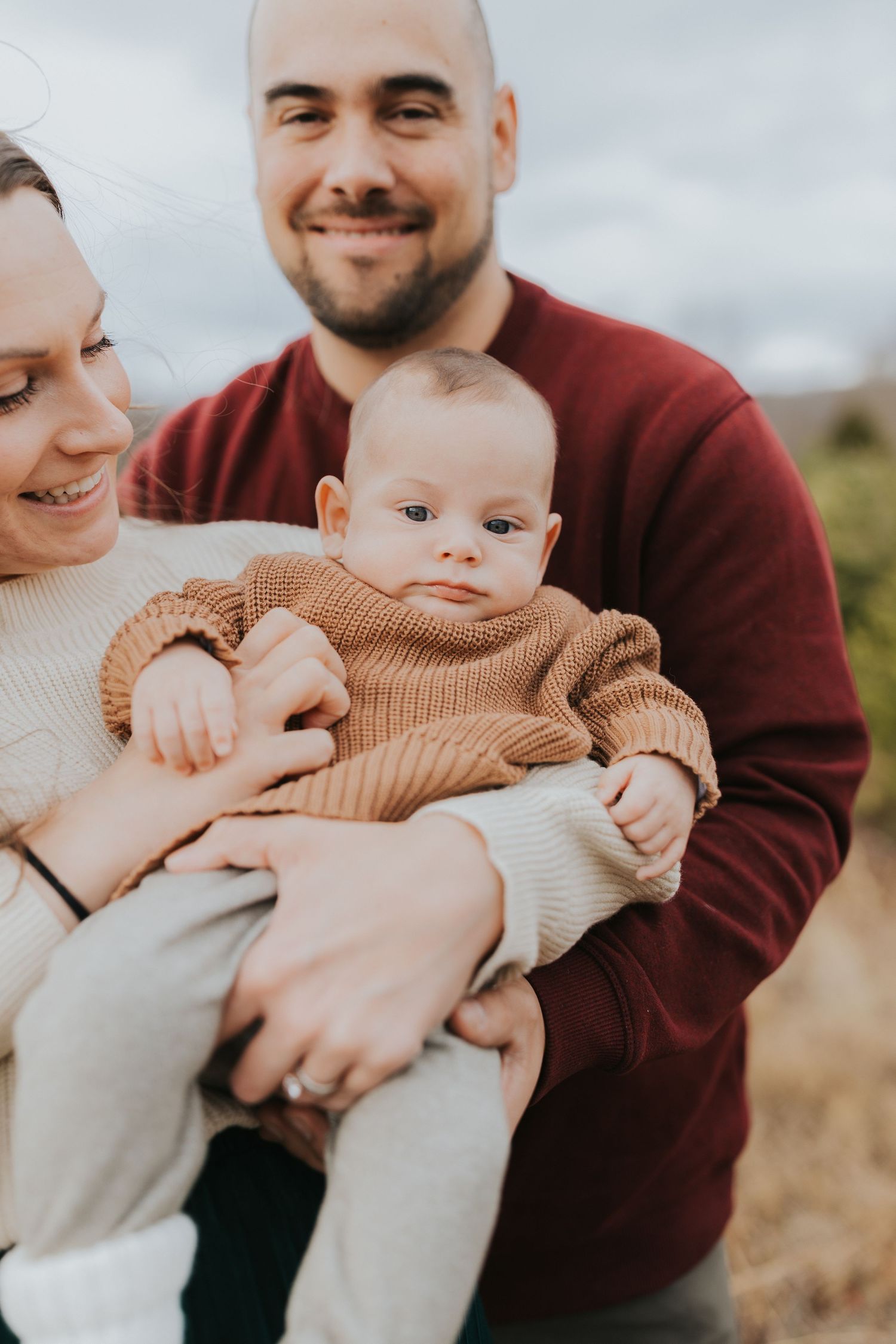 Close-up of parents holding baby at Christmas tree farm, infant looking directly at camera.