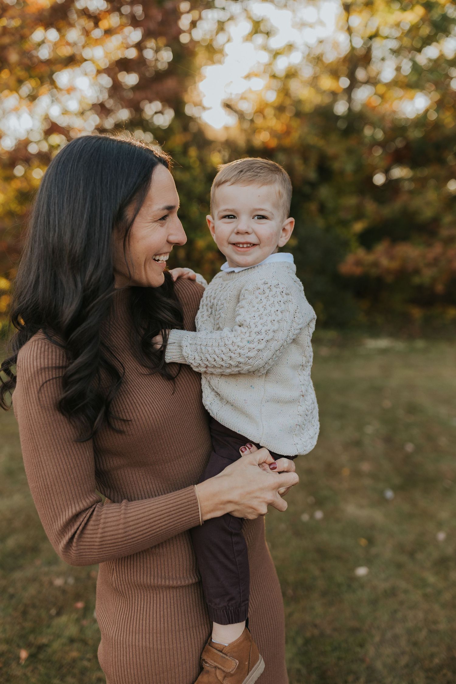 Mother in brown dress holding smiling toddler son during golden hour fall family portrait session.