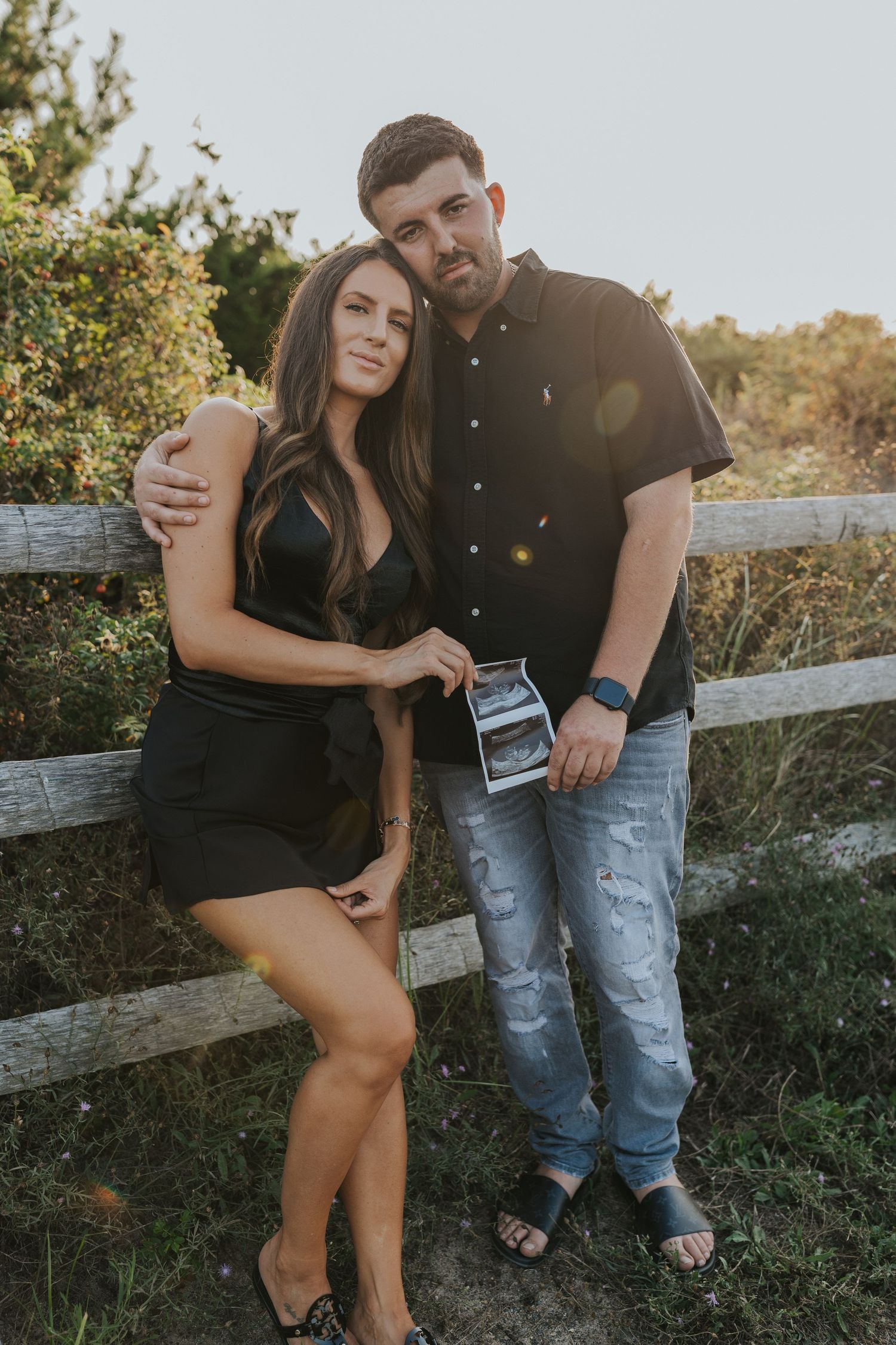 Couple posing against wooden fence holding ultrasound photos during sunset pregnancy announcement session.