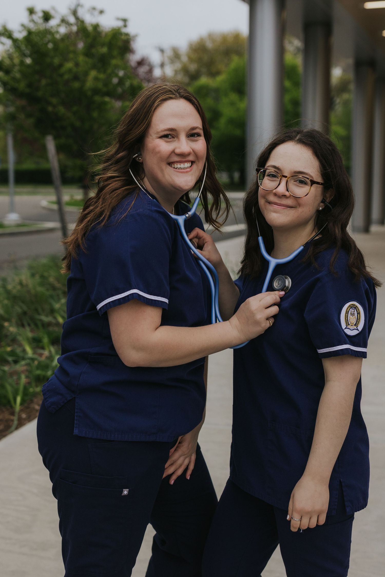 Two nursing students in navy scrubs smile while one listens with a stethoscope outdoors.