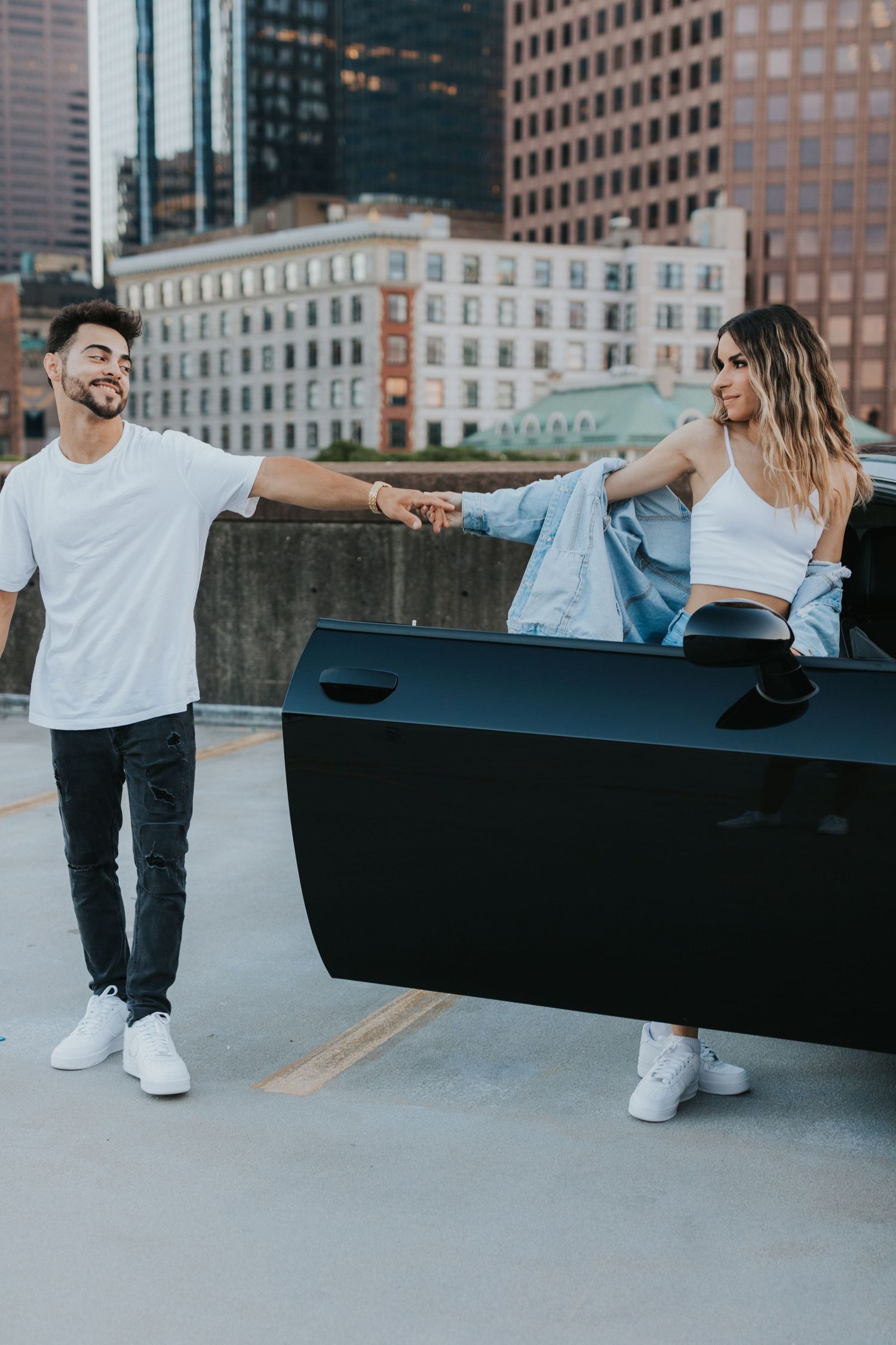 Man helping woman out of a black sports car on a rooftop parking deck with city buildings behind them.
