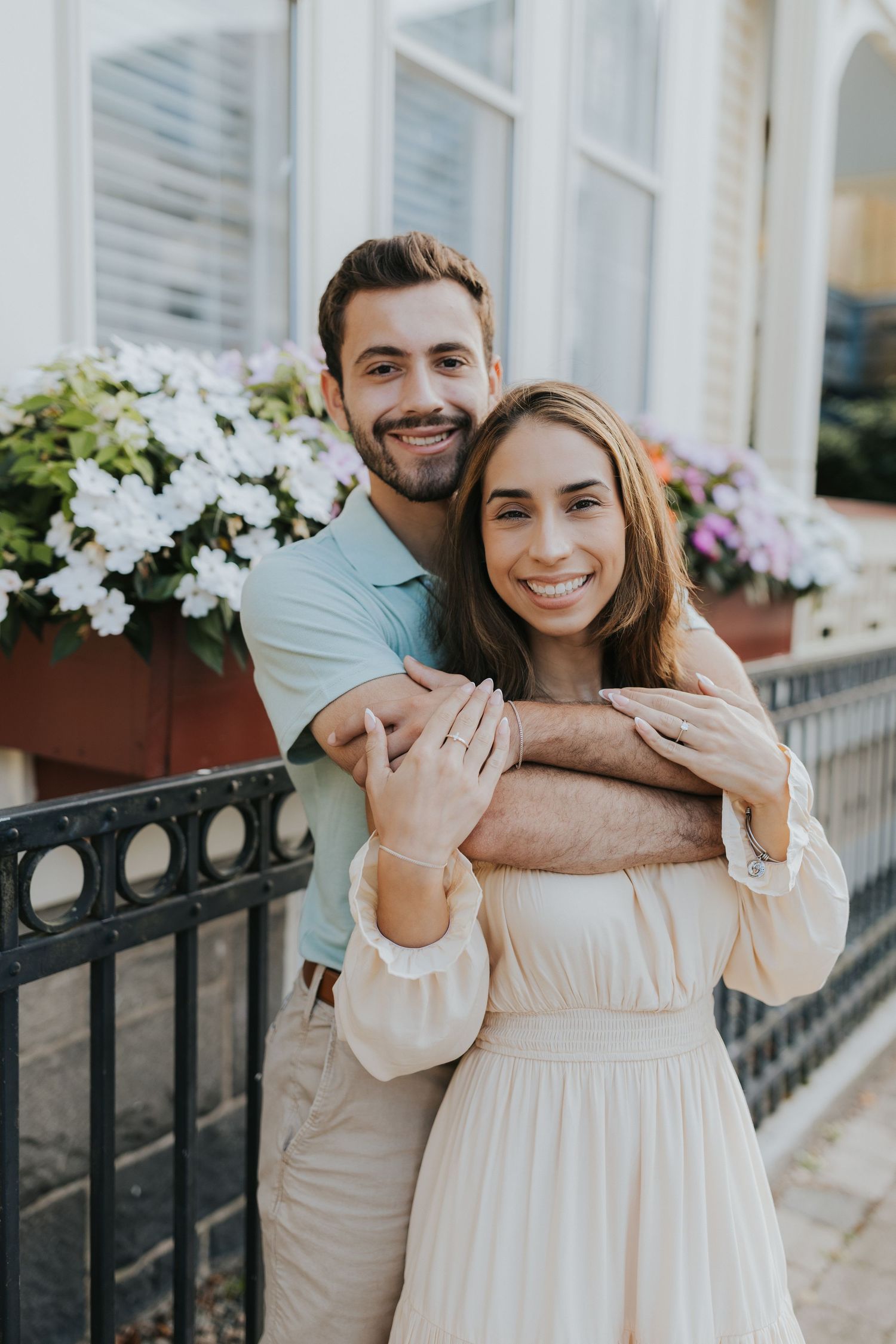 Smiling engaged couple embracing by iron fence with white flowers, woman showing engagement ring in cream off-shoulder dress.