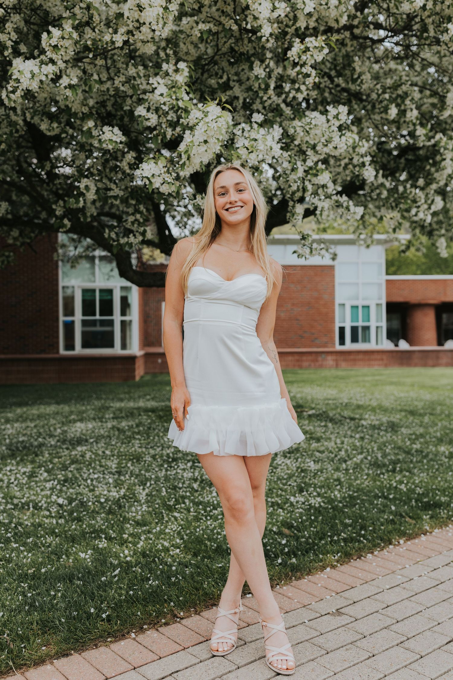Smiling blonde graduate in white strapless dress stands beneath blooming white flowering tree.
