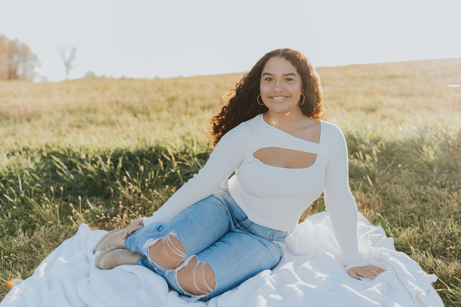 Young woman with curly hair smiling in white cutout top and ripped jeans on blanket in sunny field.
