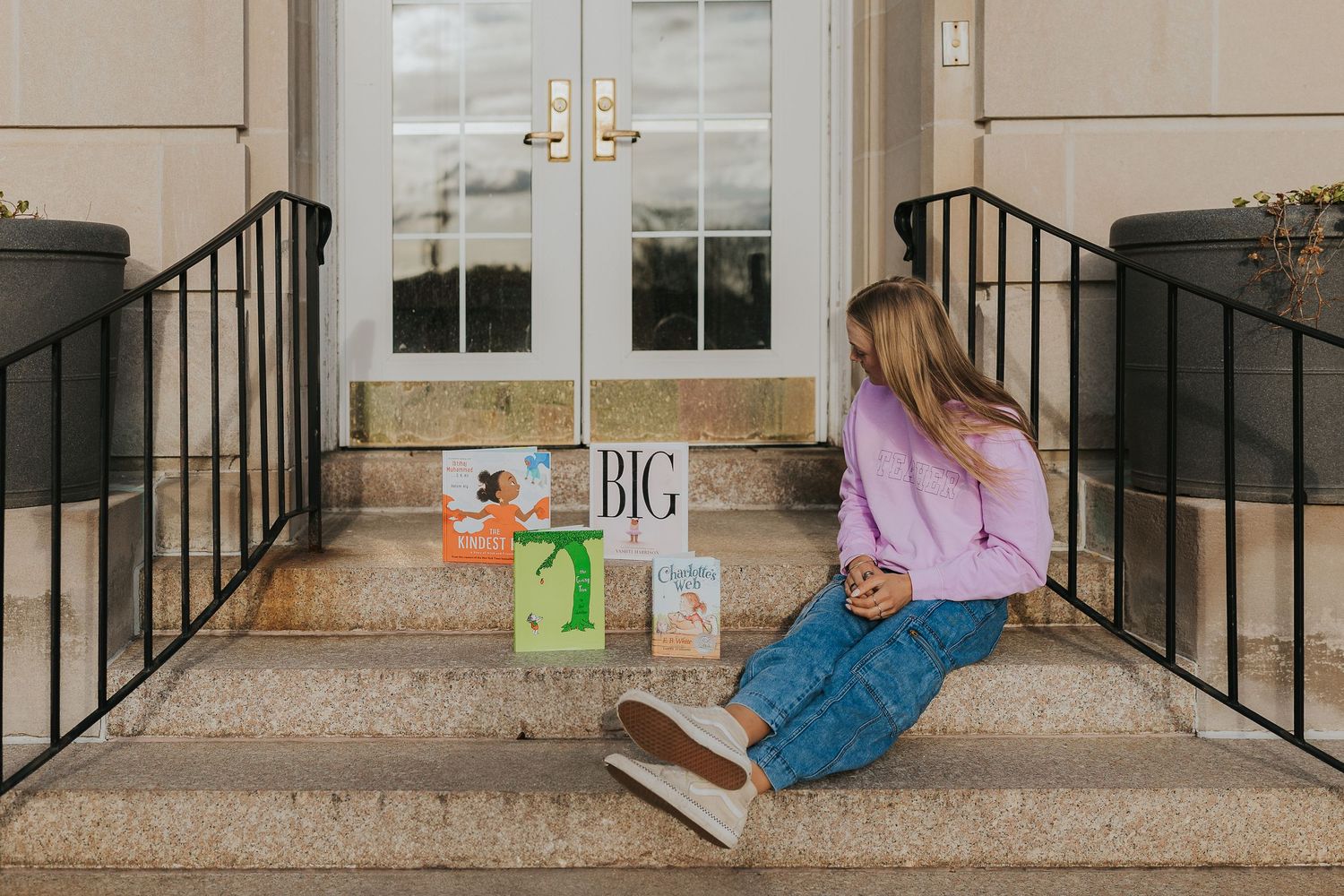 Girl sitting on brick steps surrounded by colorful childrens books outside building entrance.