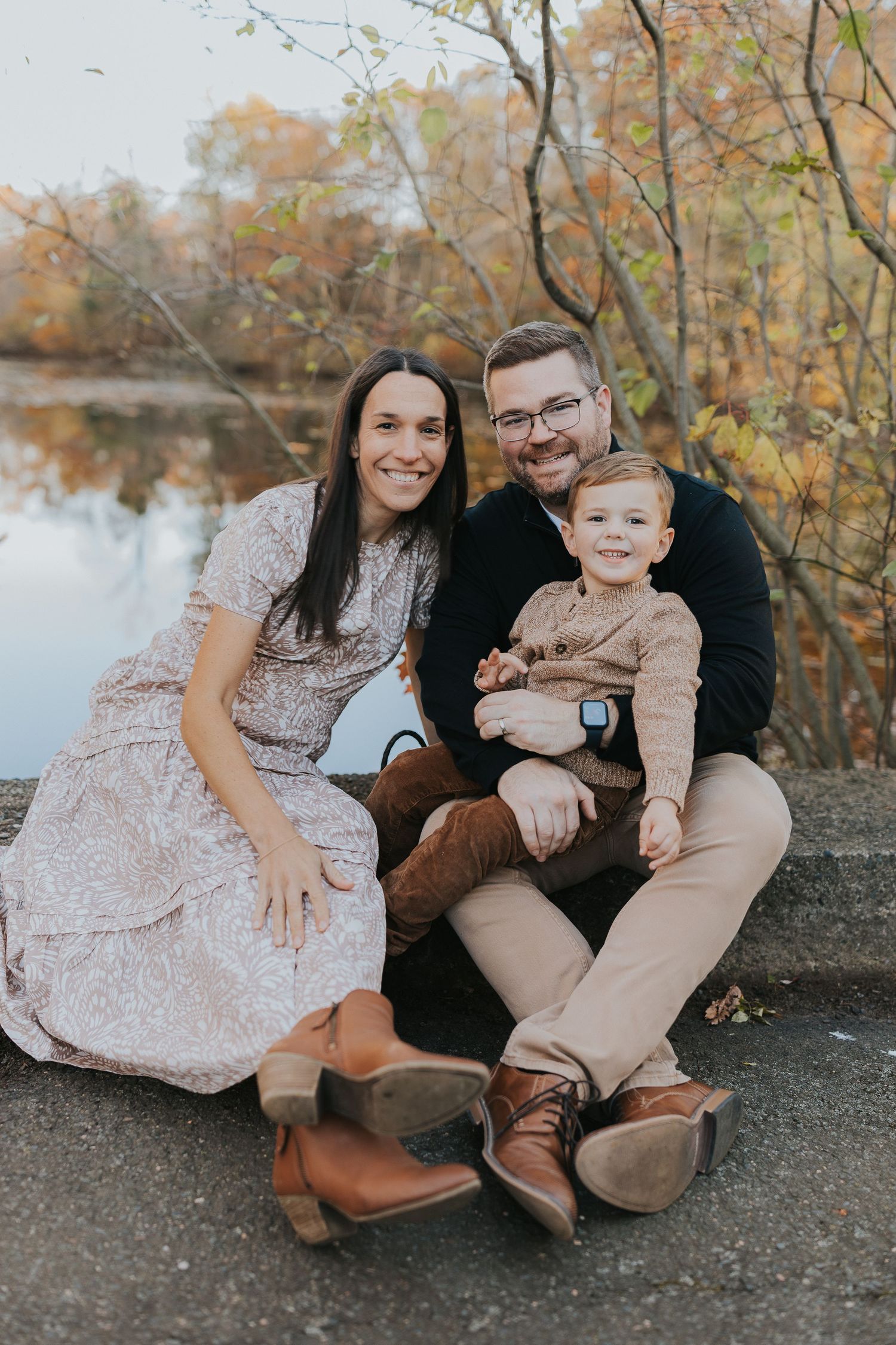 A happy family of three poses together near a reflective pond surrounded by vibrant fall foliage.