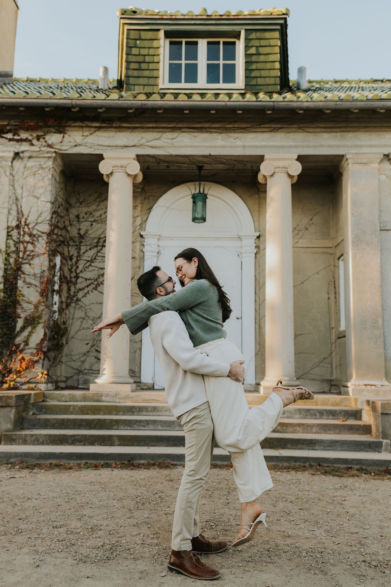 Man lifting woman joyfully in front of elegant historic building with white columns outdoors.