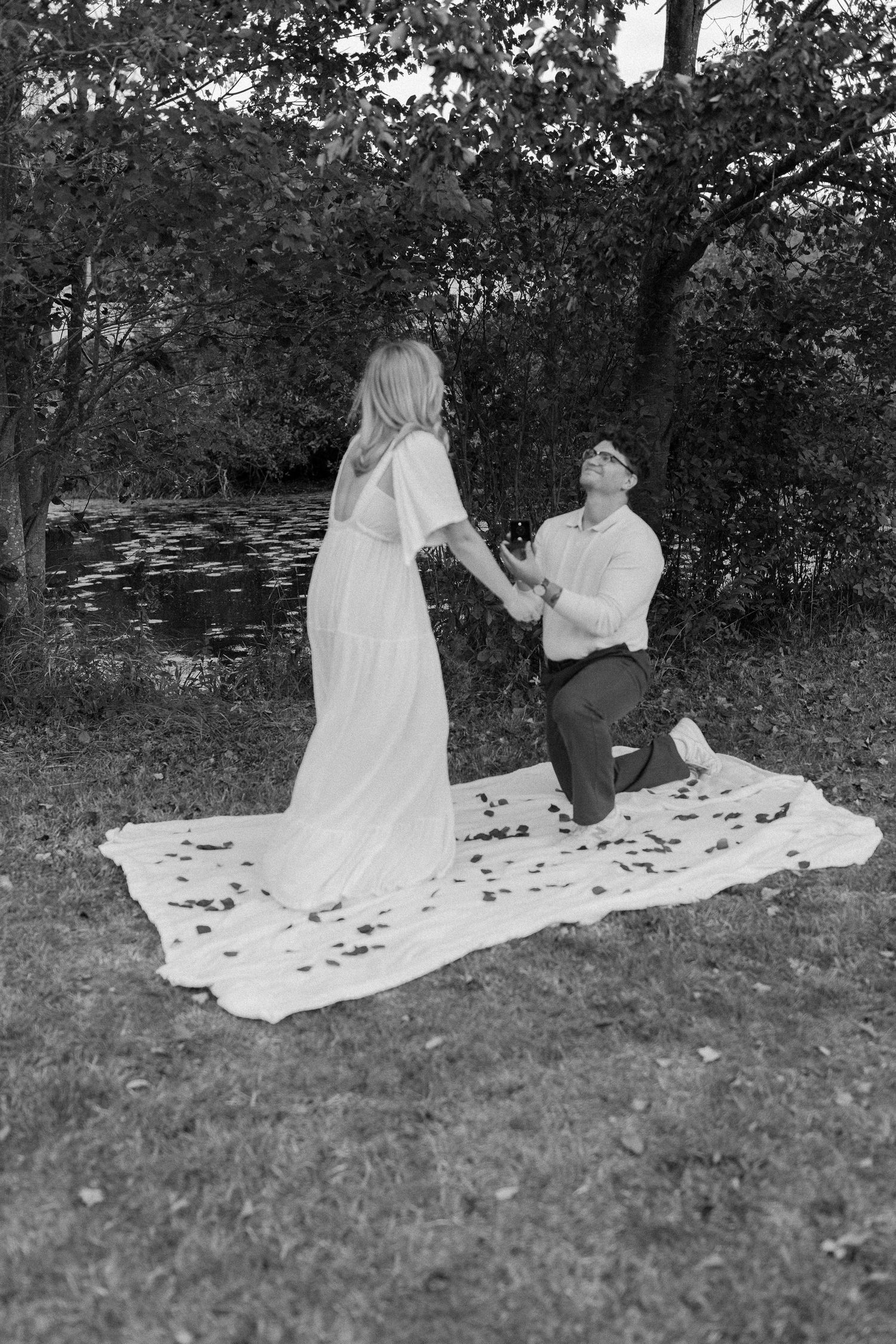Black and white photo of man proposing on one knee to woman on blanket outdoors by pond.